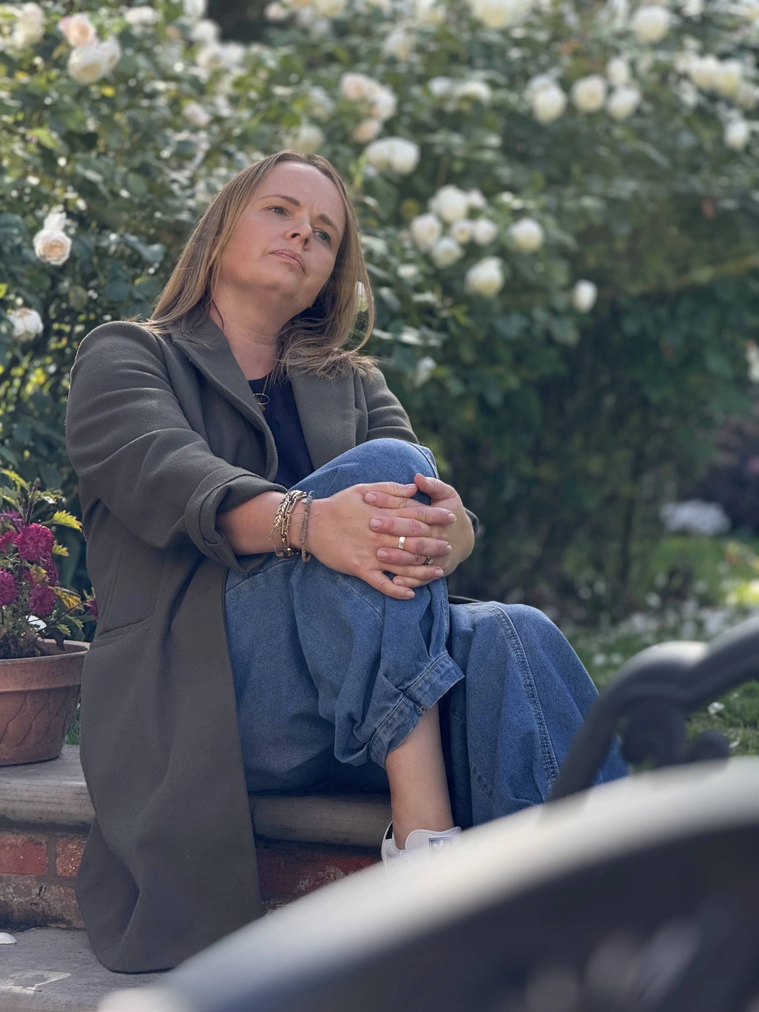 A woman sitting on a brick ledge outdoors in front of bush with white roses, wearing a dark jacket, jeans, and white sneakers, with her arms crossed over her knees and a contemplative expression.
