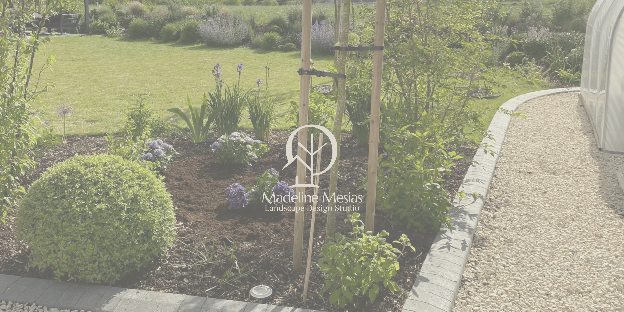 A garden bed with shrubs and flowering plants, bordered by a stone edge, with a gravel walkway beside it. There is a young tree supported by stakes and ties, and a greenhouse is visible on the side.