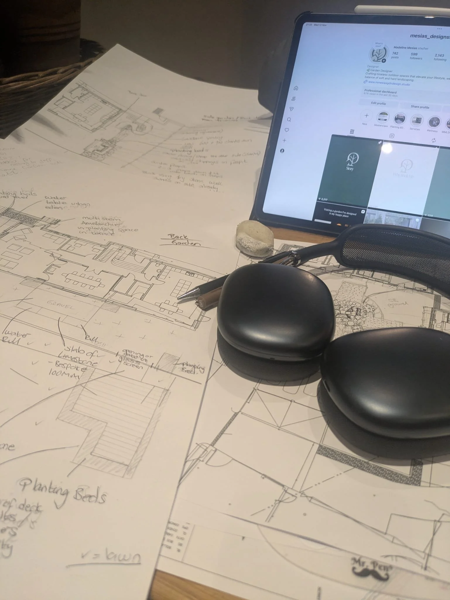 Architectural blueprints and sketches spread on a desk, with a tablet displaying a social media profile, a black pen, a pair of black earphones, and a stone on top of the blueprints.