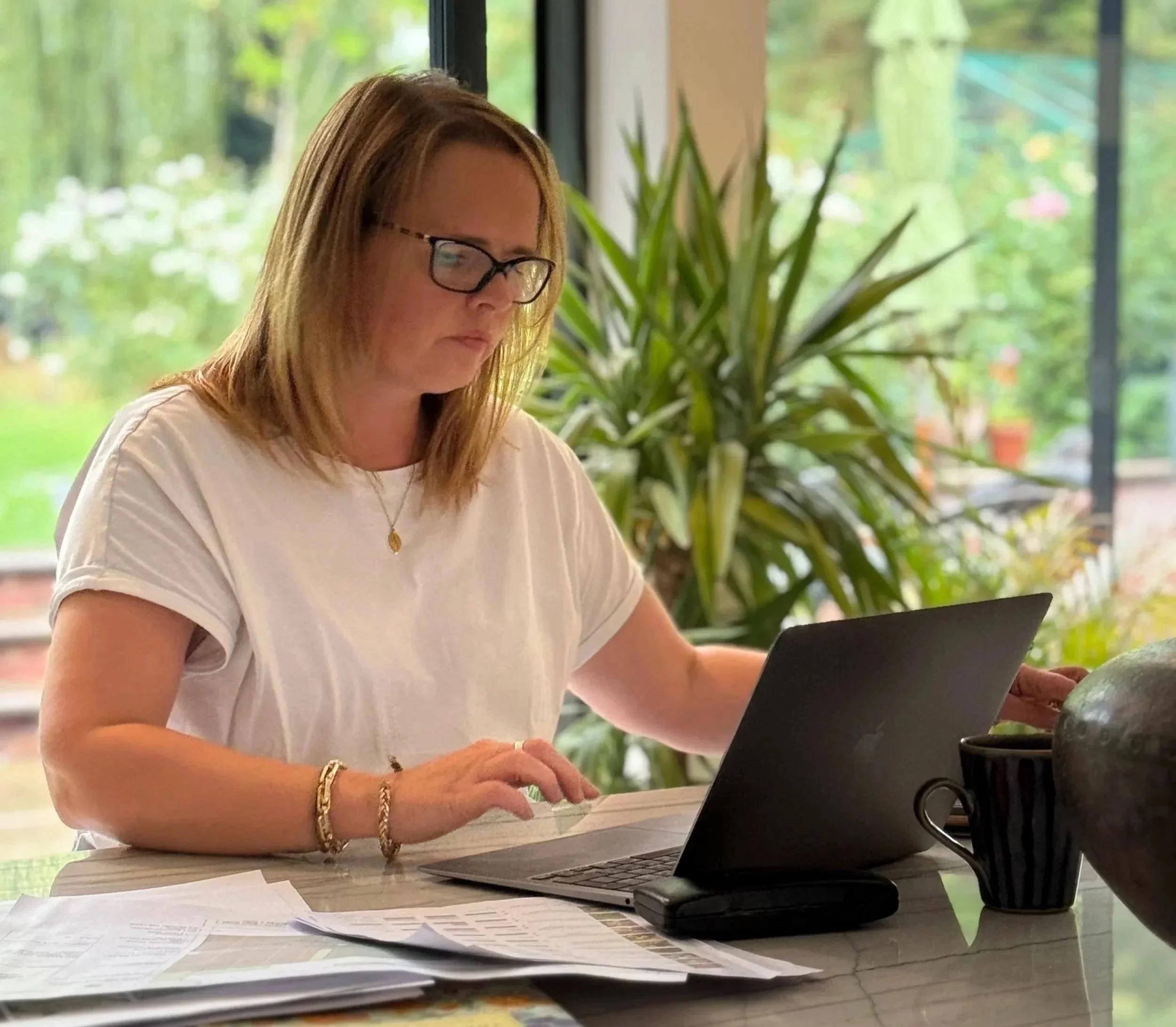 Woman with glasses working on a laptop at a table with papers, a phone, and a cup, in a bright room with large windows and outdoor greenery.