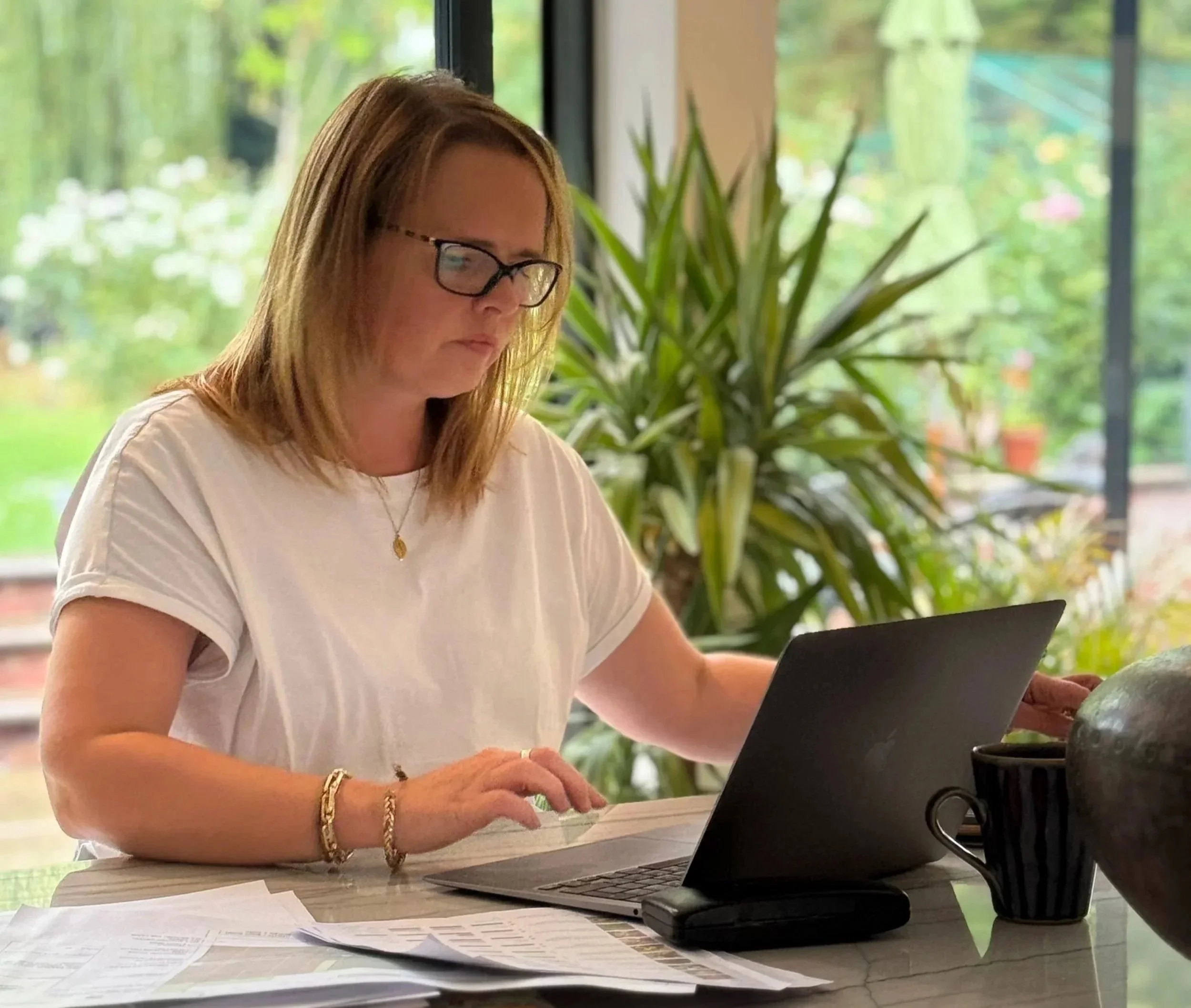 Woman with glasses working on a laptop at a desk with papers and a coffee mug, in a room with large windows and green plants outside.