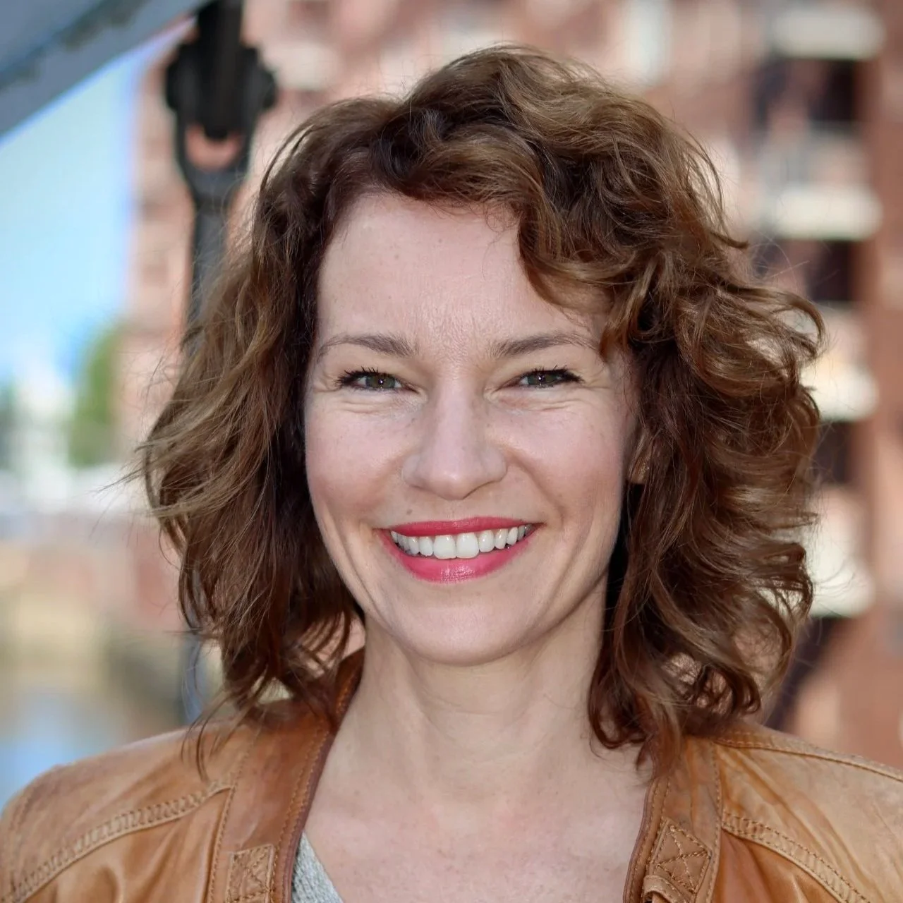 Lächelnde Frau mit lockigem, rötlichem Haar vor einem urbanen Hintergrund.