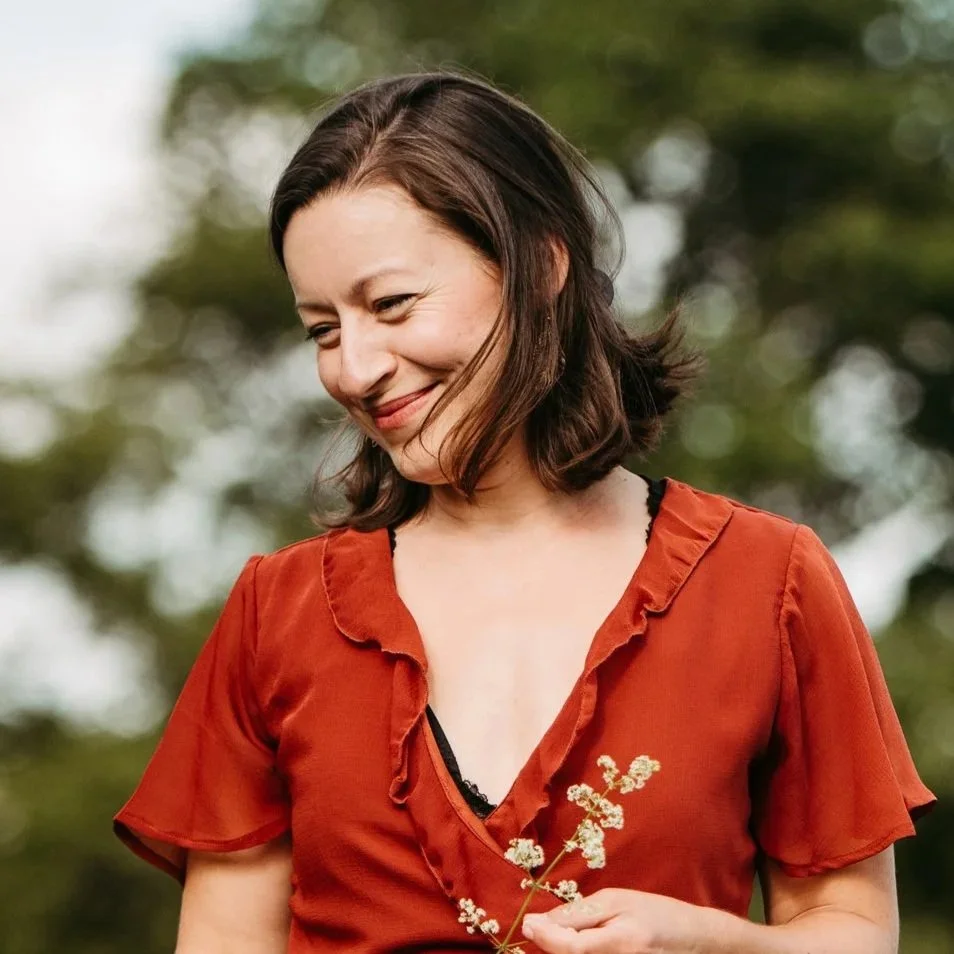 Eine lachende Frau mit dunklem, schulterlangem Haar trägt ein rotes Kleid und hält eine kleine Blumenstängel in der Hand, im Hintergrund sind Out-of-Fokus-Bäume.
