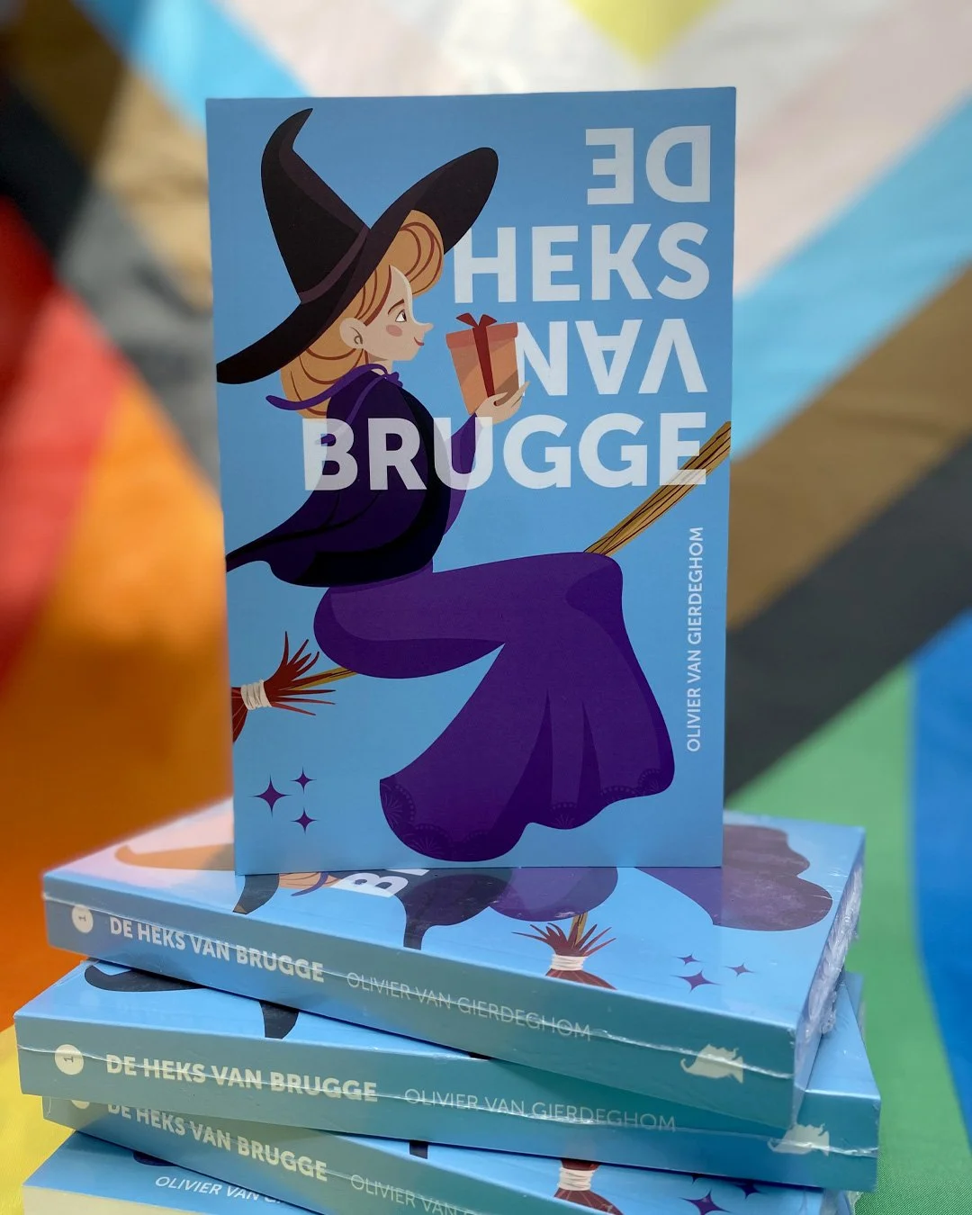 Kinderboek met een illustratie van een heks op de cover, zittend op een bezem en een cadeau in haar hand, met de titel 'De Heks Van Brugge' en de naam van de auteur Olivier Van Gierdeghom.