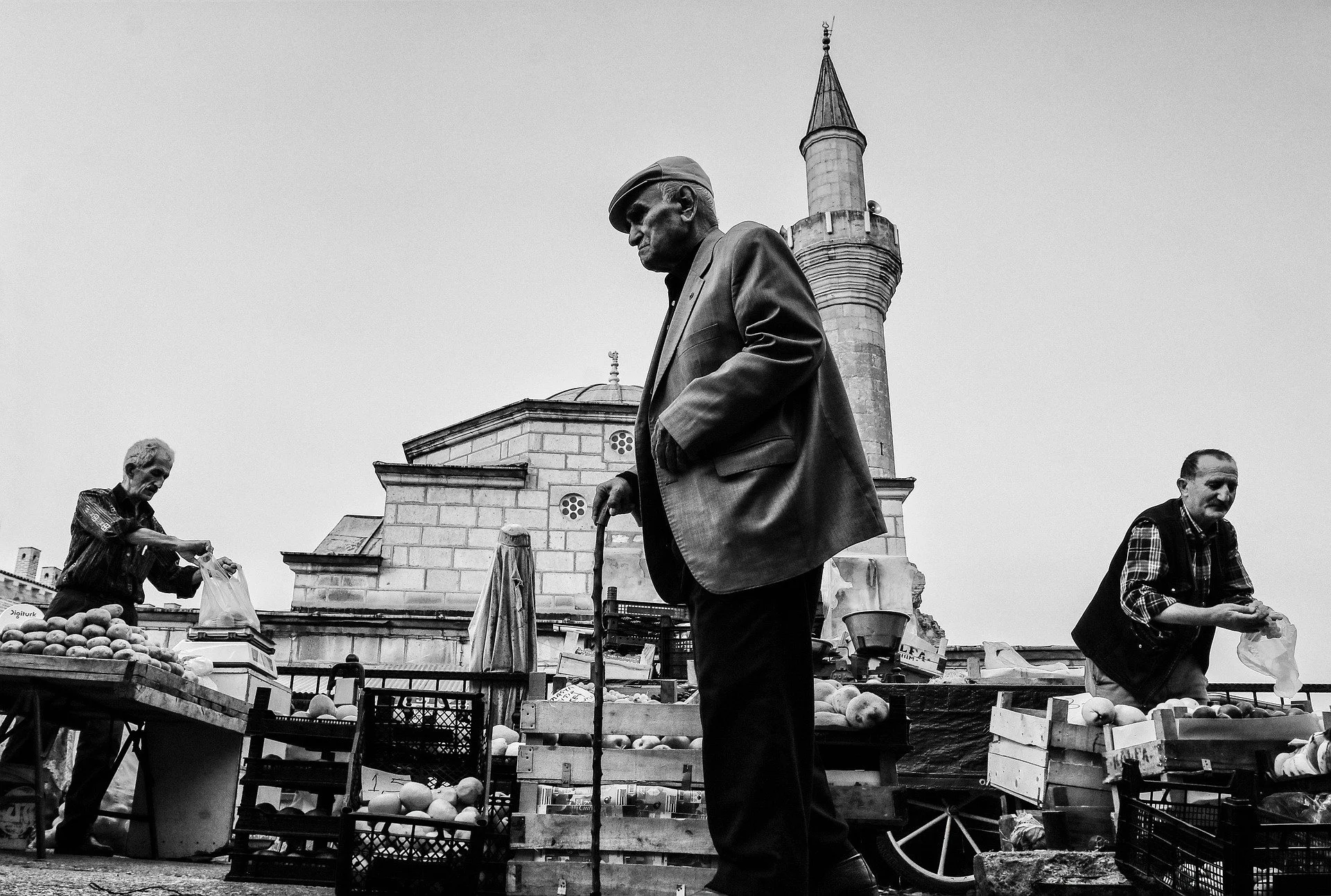 Uomo anziano con cappello e bastone davanti a un mercato di frutta, con un edificio storico in background.