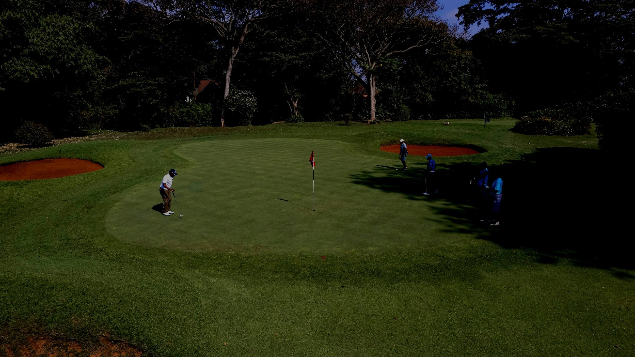 A group of people playing golf on a golf course, with a golfer preparing to putt on the green next to a flagstick, and others nearby watching or waiting.