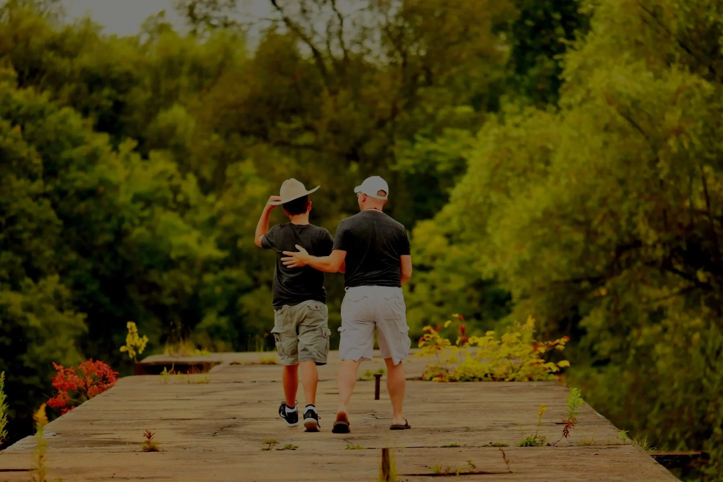 Two people walking on a wooden bridge surrounded by trees, one with a hat and the other with a cap, with their arms around each other.