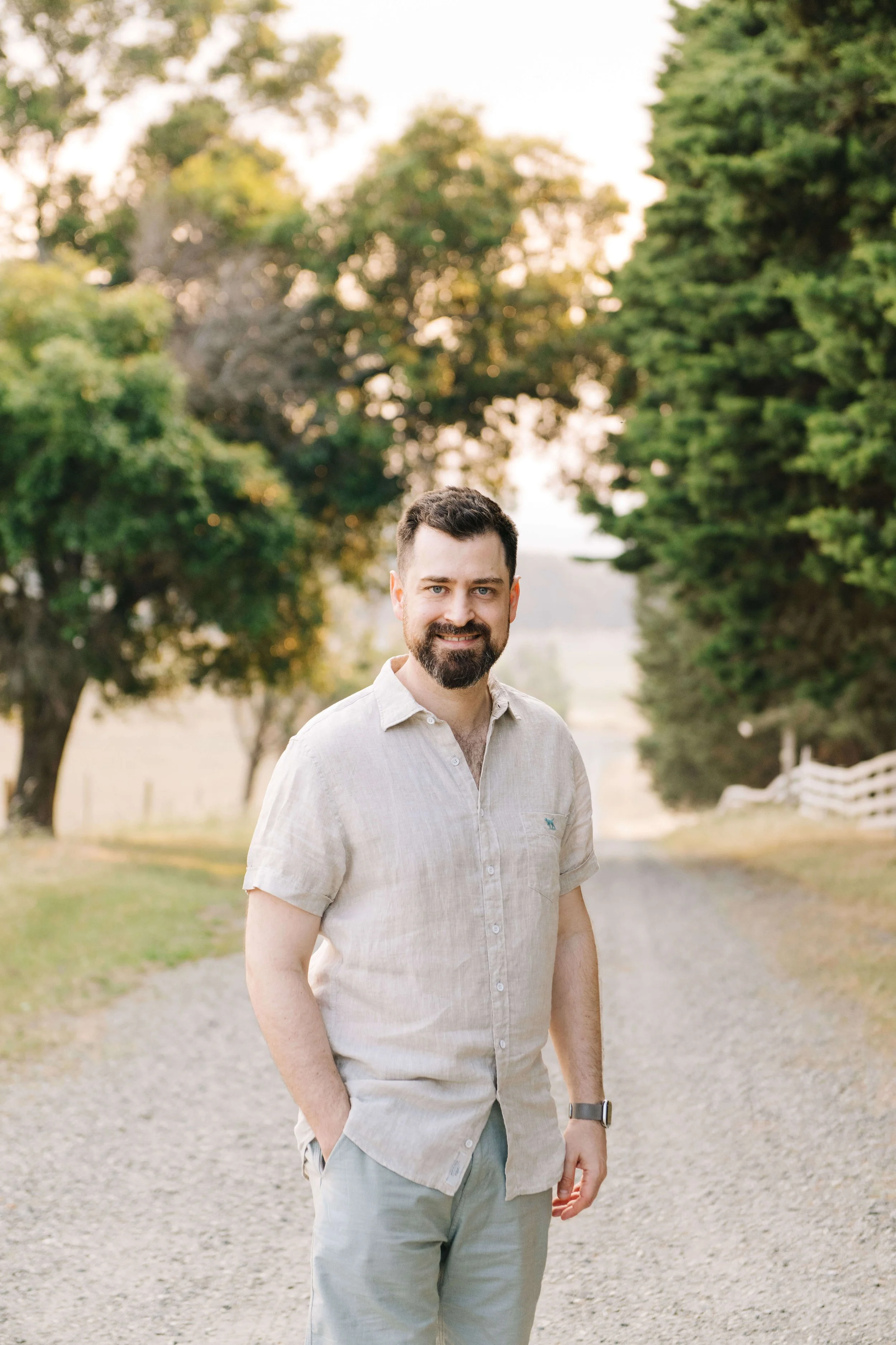 A young man with a beard and short dark hair standing outdoors on a gravel path lined with trees. He is wearing a light-colored short-sleeve button-up shirt and light-colored pants, with his left hand in his pocket. The background features green trees and a white fence, with soft, warm lighting suggesting late afternoon or early evening.