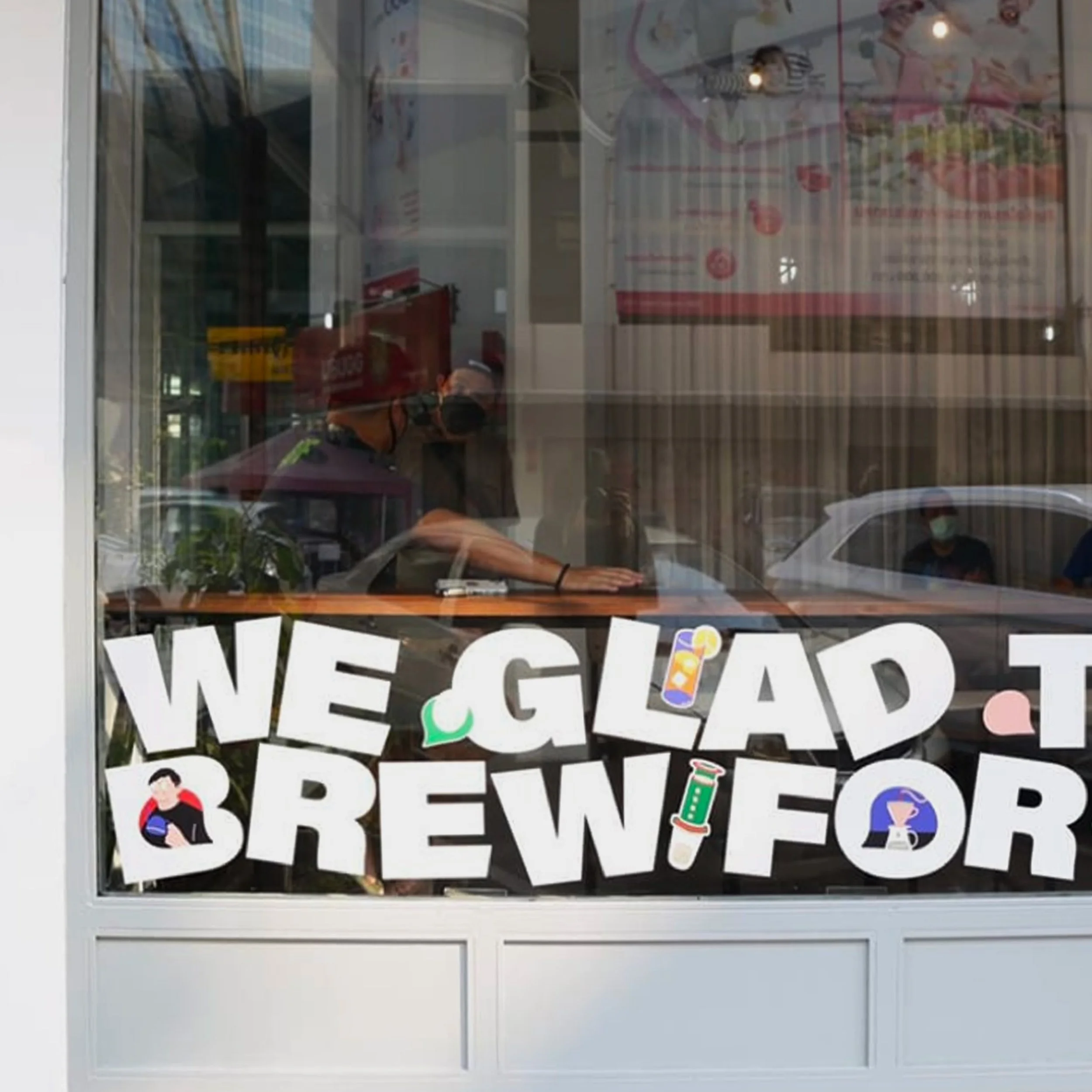 A window sign that reads 'We Glad to Brew for' with colorful illustrations of a lemon, a cocktail glass, a syringe, and a person wearing a hoodie.