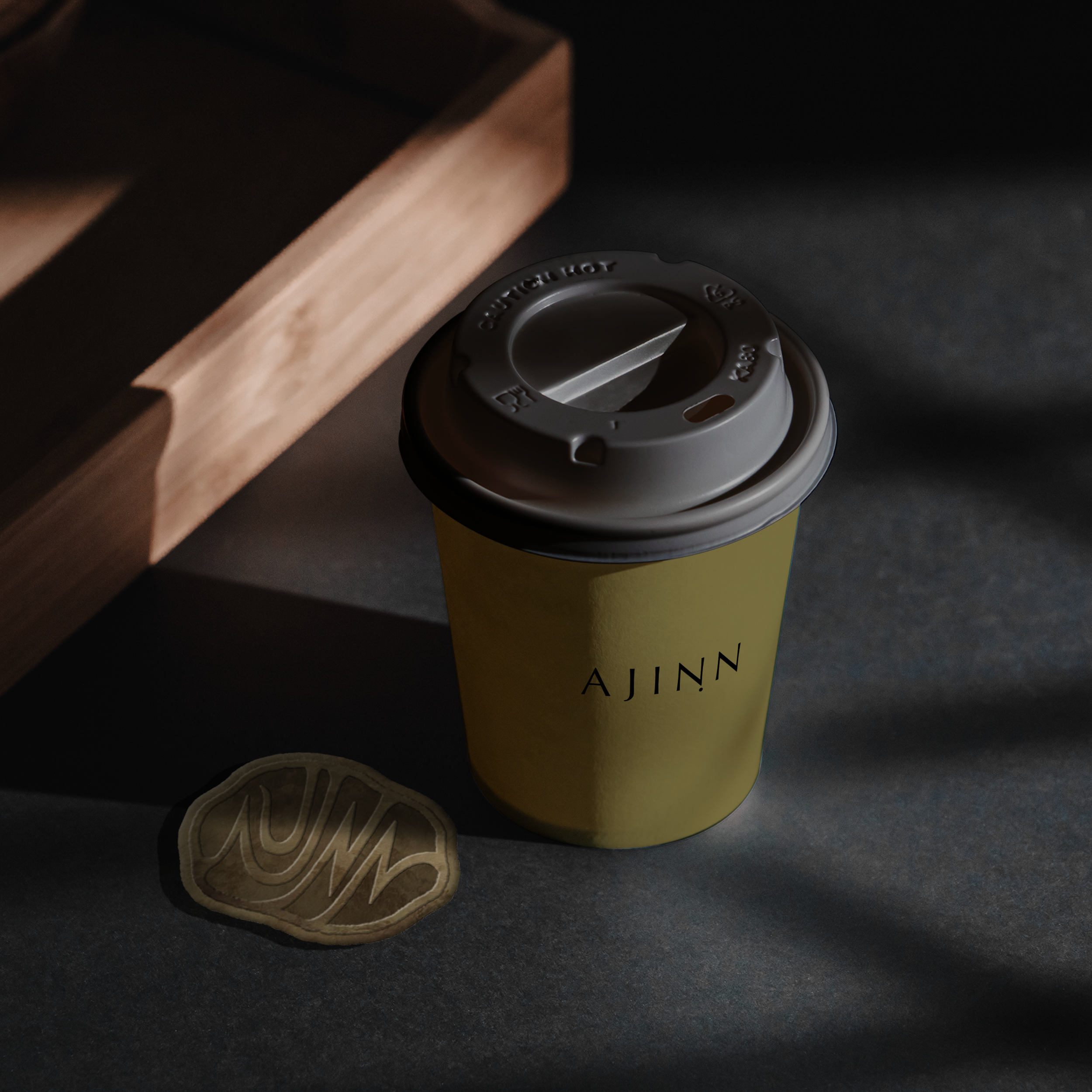 A yellow paper coffee cup with a plastic lid reading 'AJINN' on a dark surface, with a cookie nearby and a wooden object casting shadows.