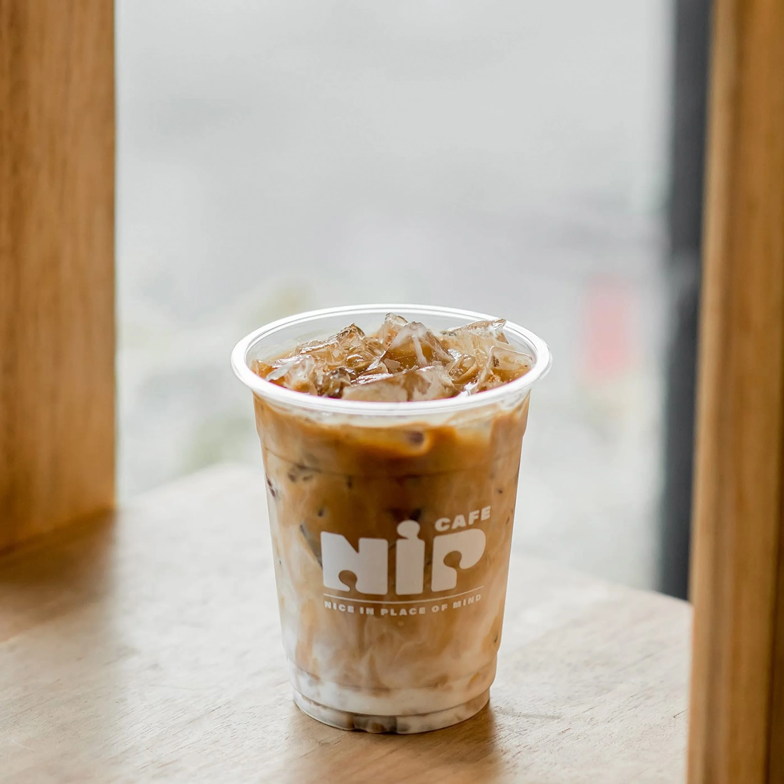 Iced coffee in a clear plastic cup with a logo on it, placed on a wooden surface near a window with wooden framework.