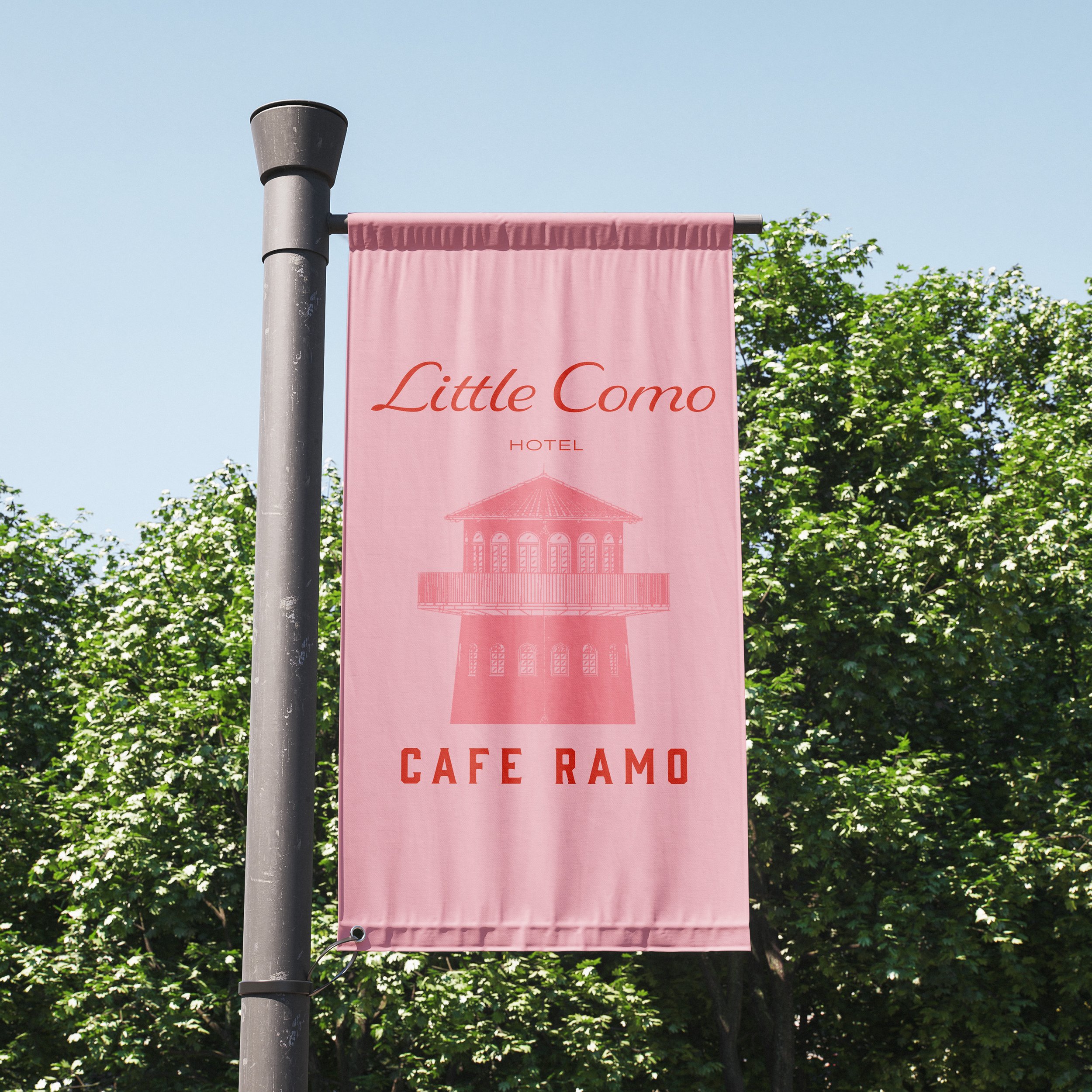 A pink vertical banner for Little Como Hotel Cafe Ramo hanging on a street pole with green trees in the background.