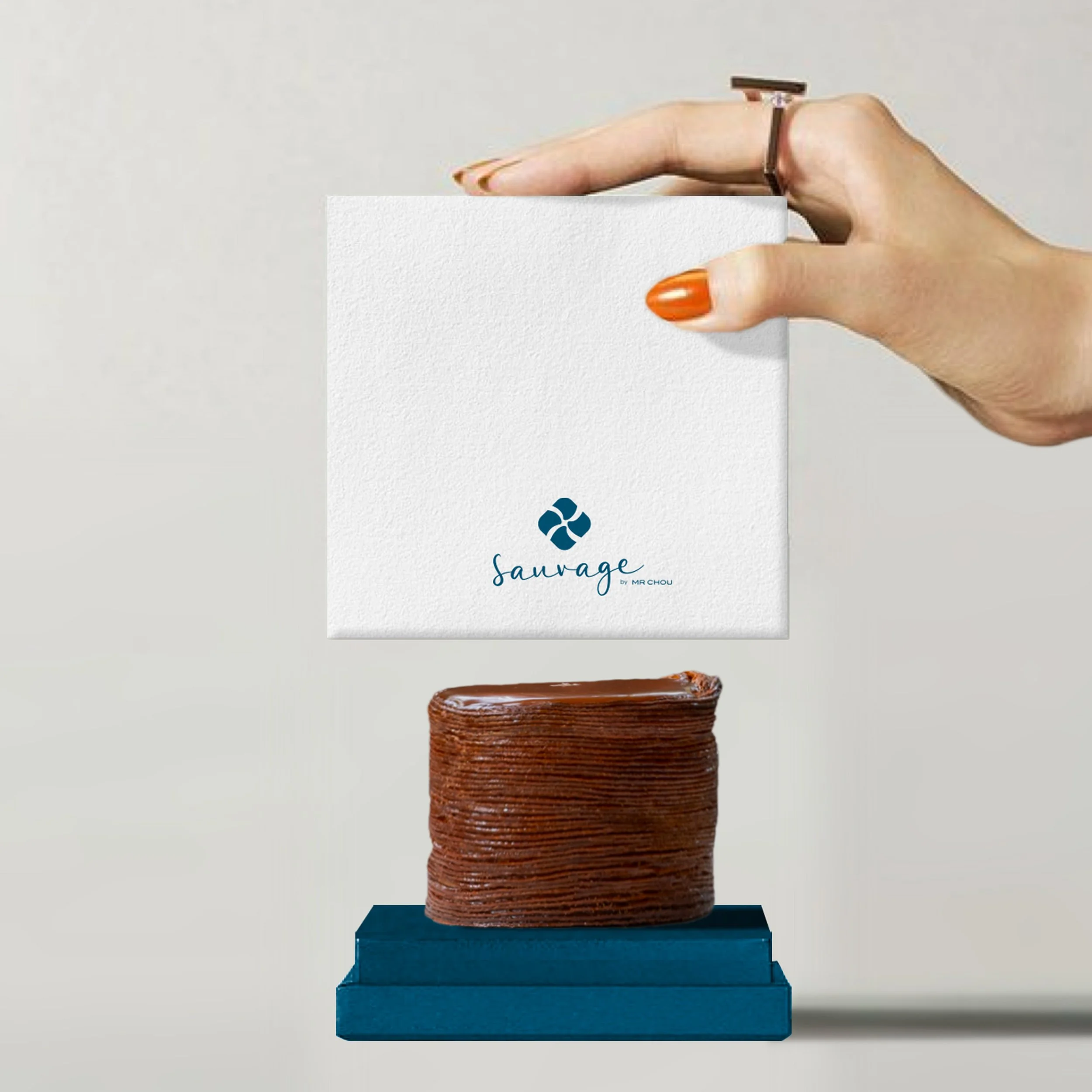 A hand with orange nail polish and a ring holds a white card with a blue logo and the text 'sauvage' above a chocolate layered cake on a blue stand.