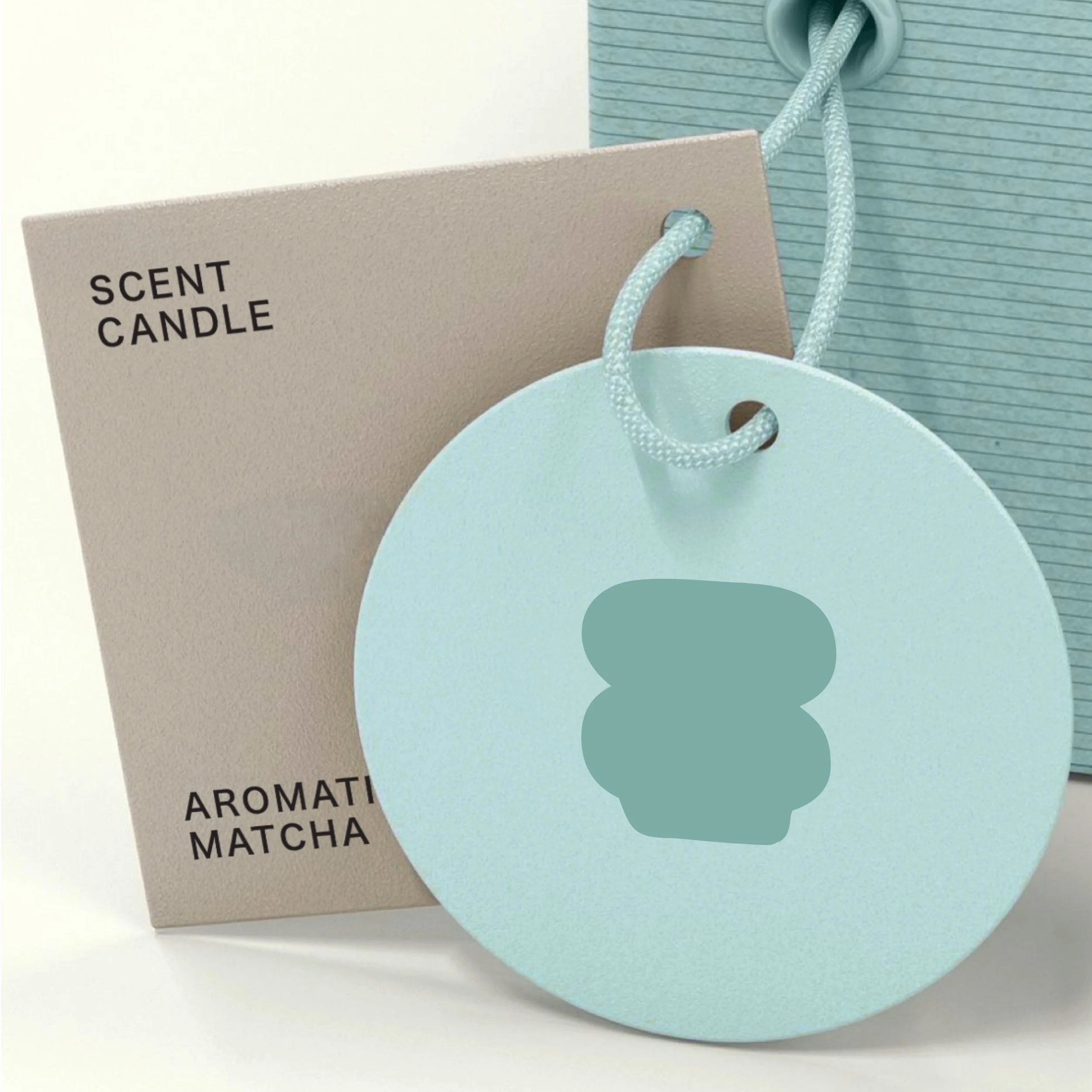 A light blue round aromatherapy matcha scented candle with a hole at the top attached to a white rope handle, placed in front of a beige box labeled "SCENT CANDLE" and "AROMATIC MATCHA."
