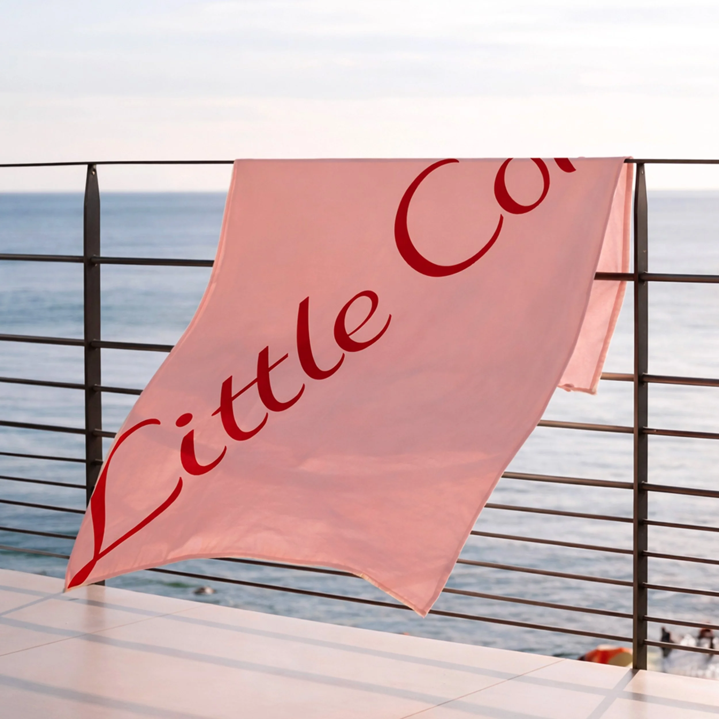 Pink towel with the words 'LITTLE CO' hanging on a deck railing overlooking the ocean.
