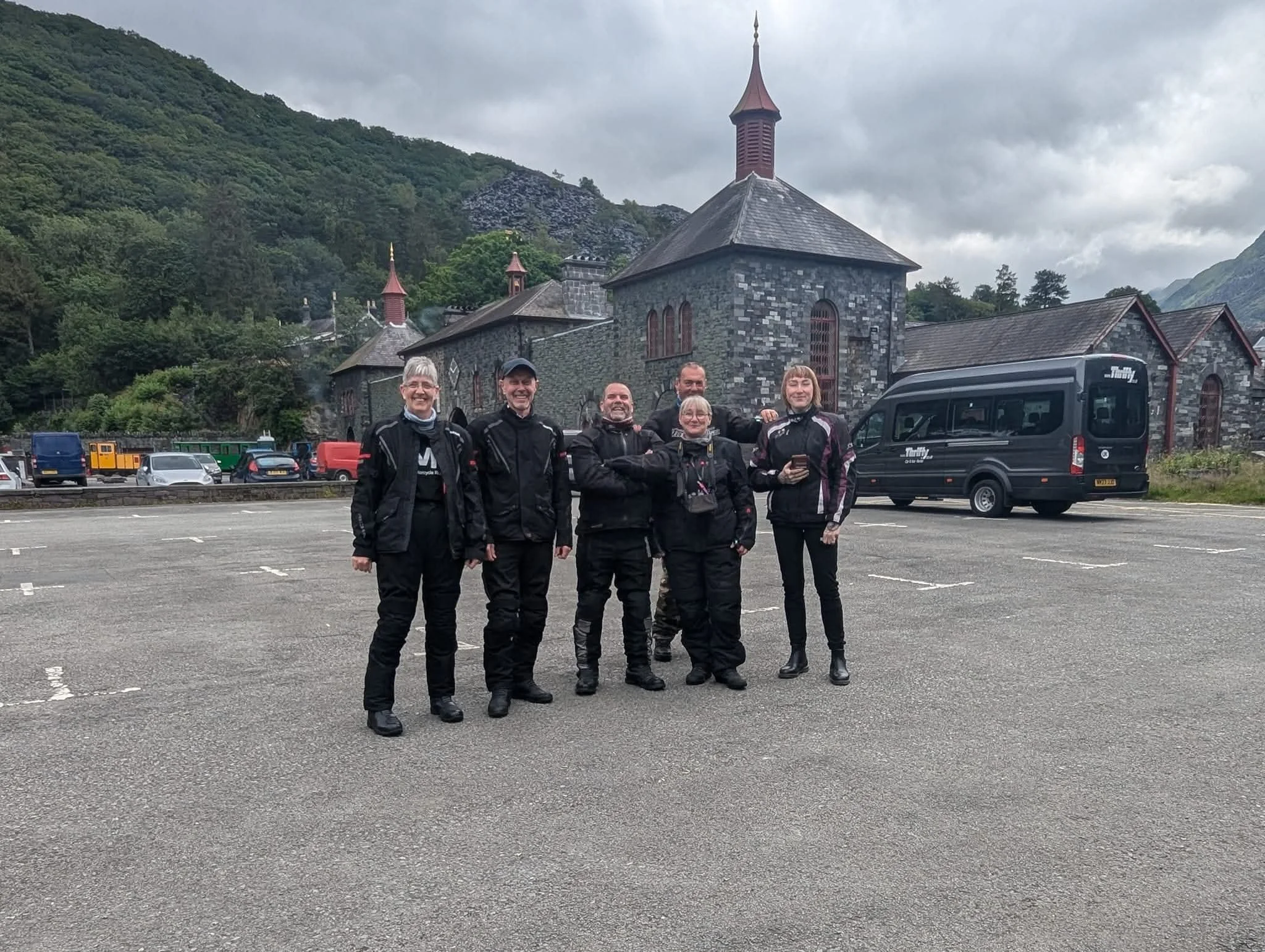 Group of six people in motorcycle gear standing in a parking lot with a stone church and mountainous landscape in the background.