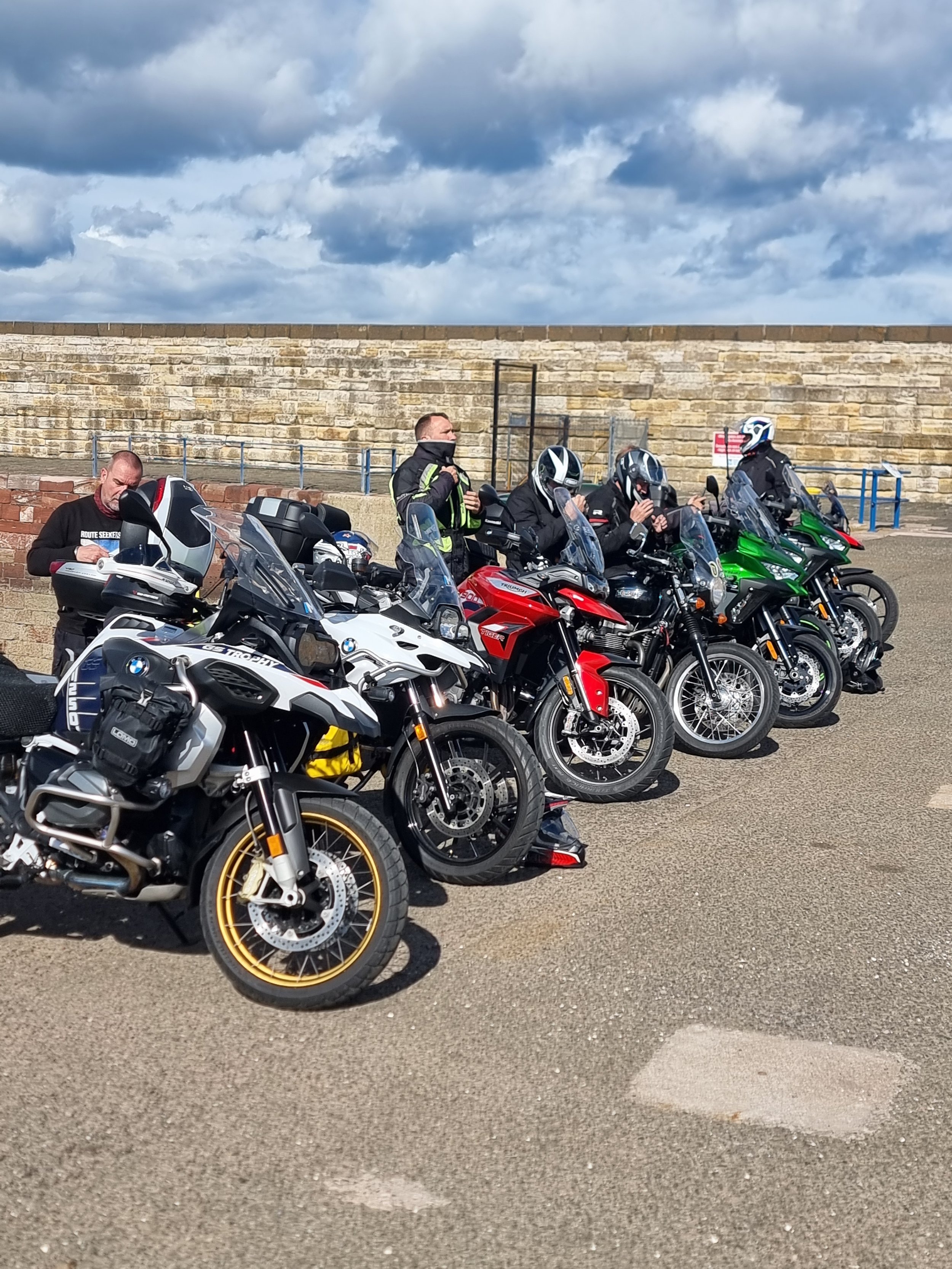 A group of six motorcyclists in gear standing and sitting next to their parked adventure motorcycles in an outdoor parking lot, with a stone wall and cloudy sky in the background.
