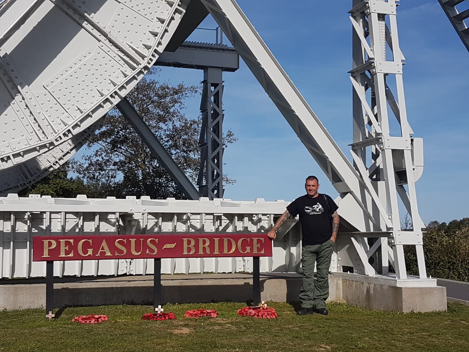 A man standing next to a sign that reads 'Pegasus Bridge' in front of a large white metal structure of a bridge with a blue sky background.