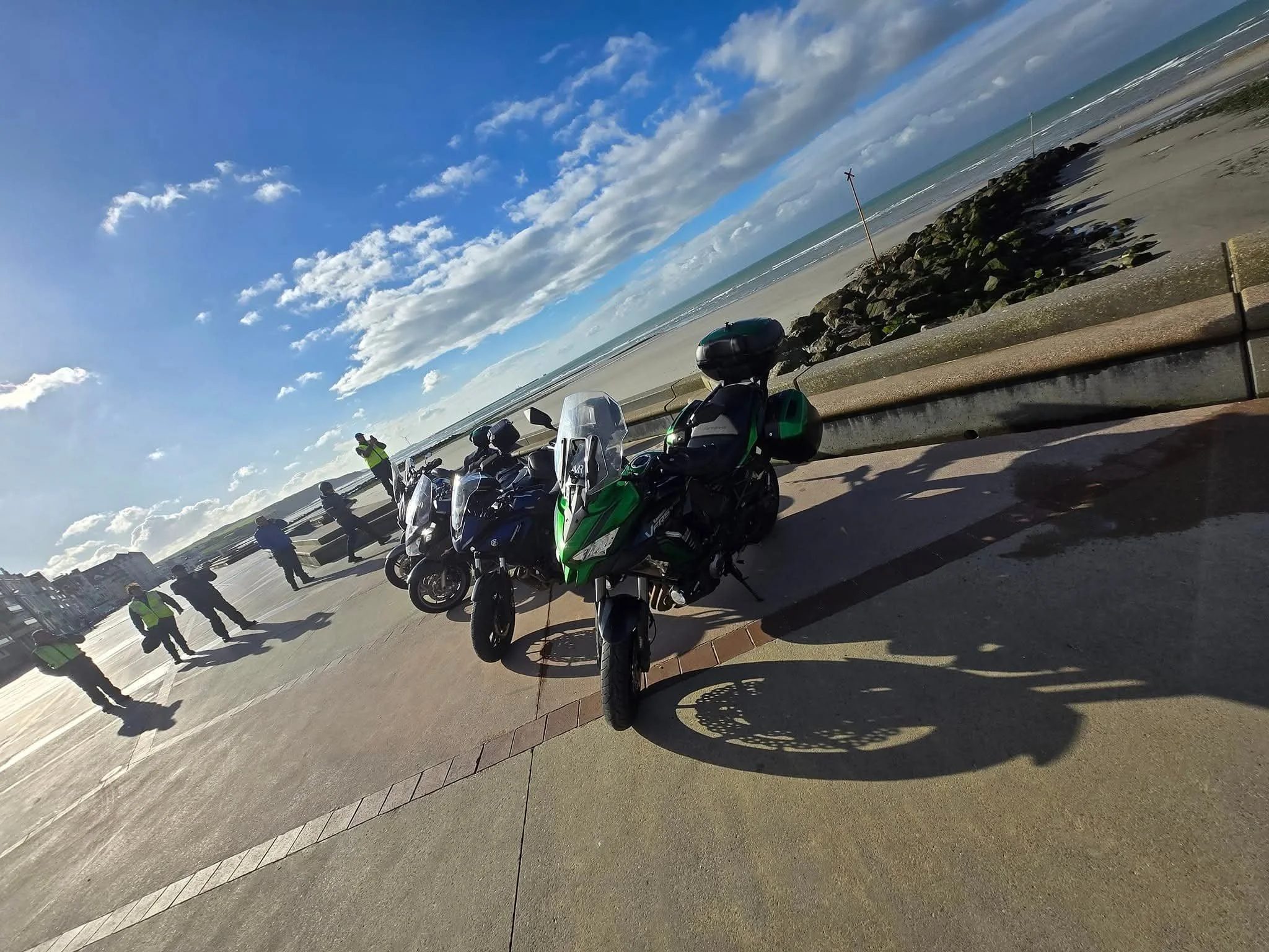 Motorcycles parked by the beach, with a concrete barrier and large rocks, people standing and talking, cloudy sky with some patches of blue.