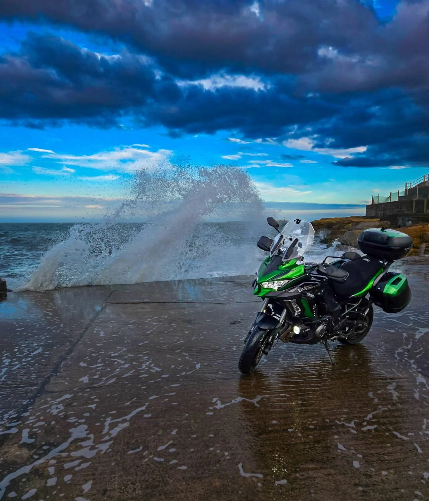 A green and black motorcycle parked on a wet concrete surface near the ocean with waves splashing against the rocks and a cloudy sky overhead.