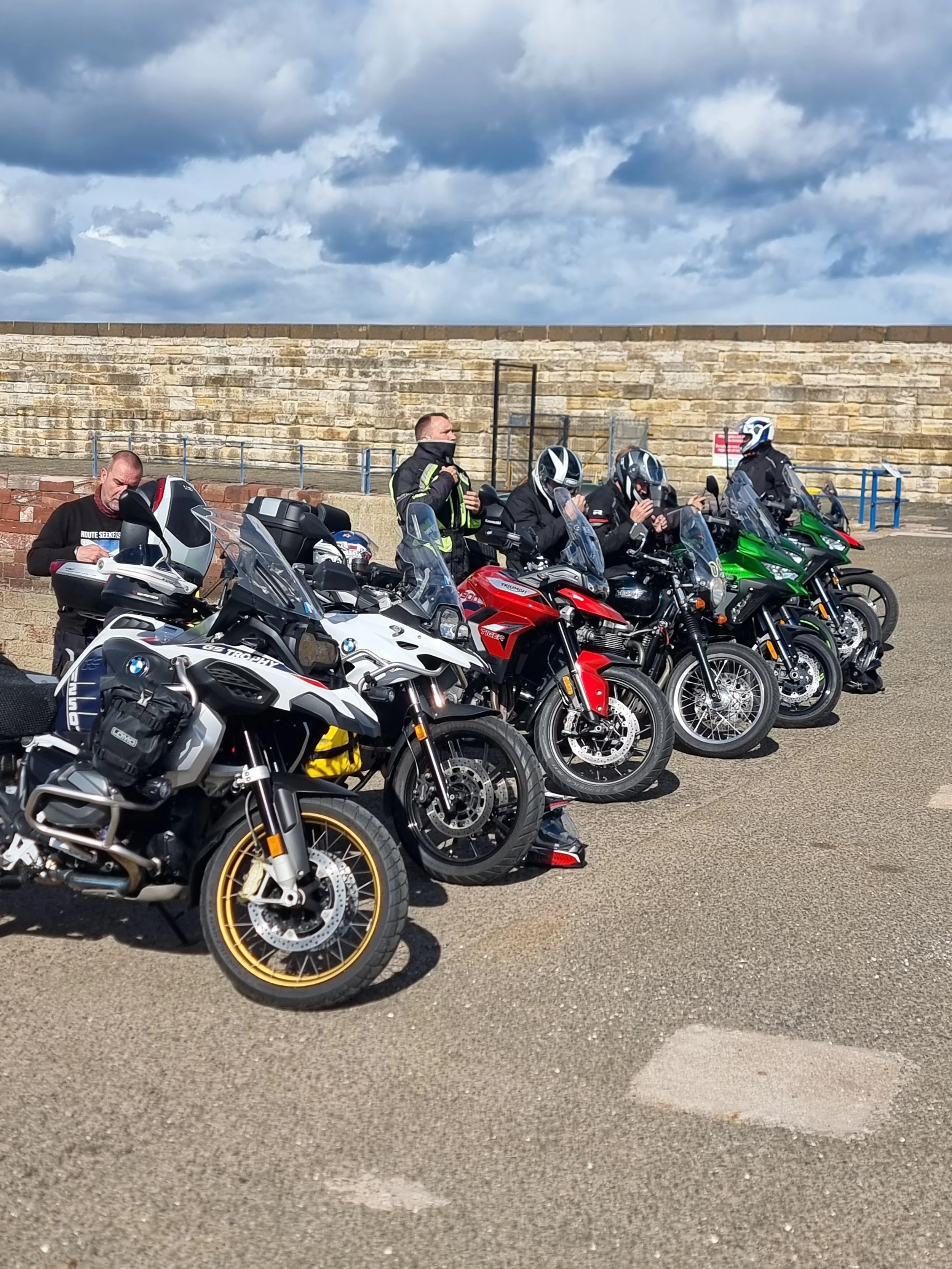 Group of six motorcyclists sitting on their bikes, preparing or resting, with a stone wall and cloudy sky in the background.