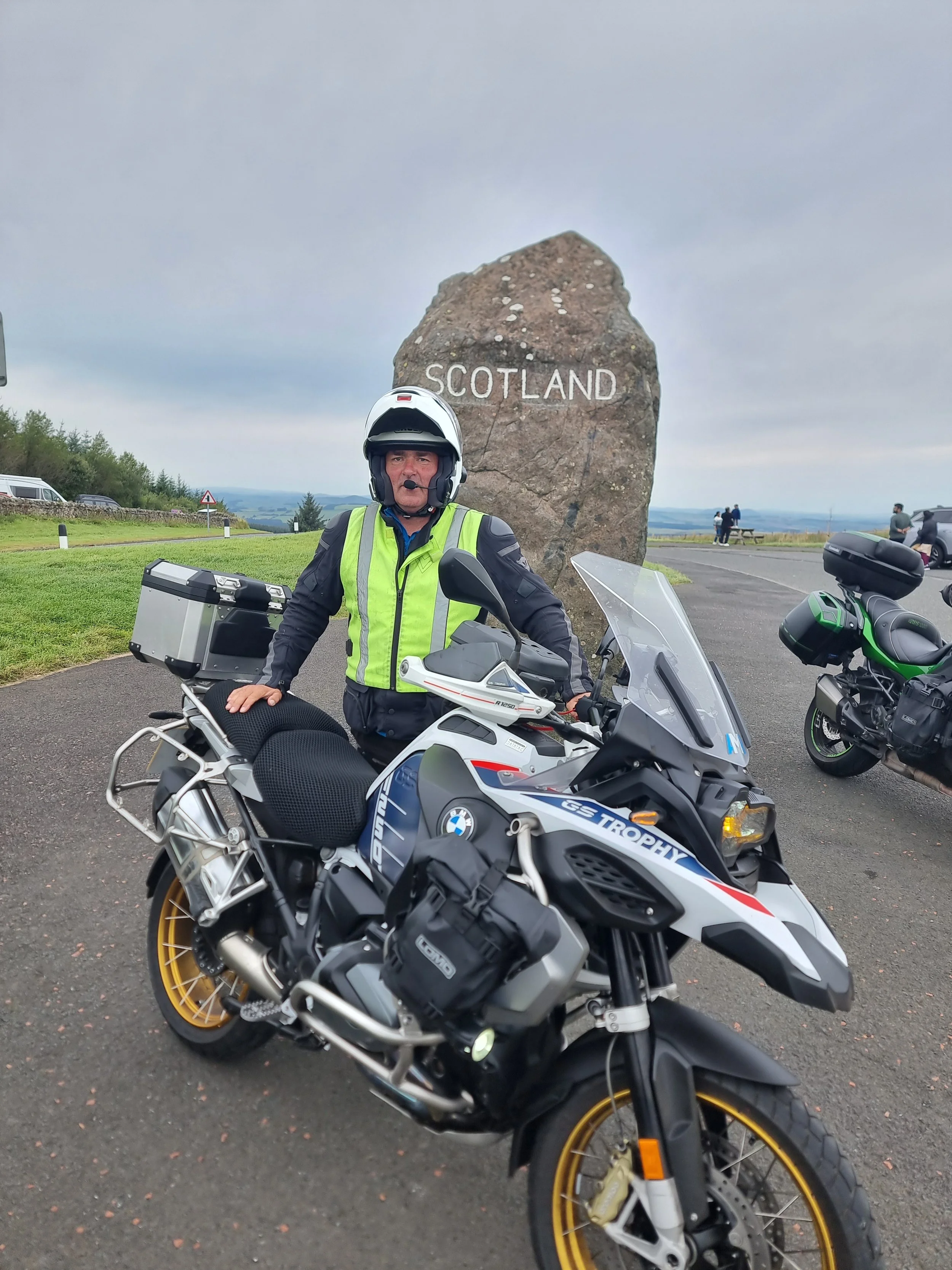 A man in a motorcycle helmet and yellow safety vest standing next to a BMW GS Trophy motorcycle at a Scotland roadside overlook, with a large rock behind him with 'Scotland' painted on it.
