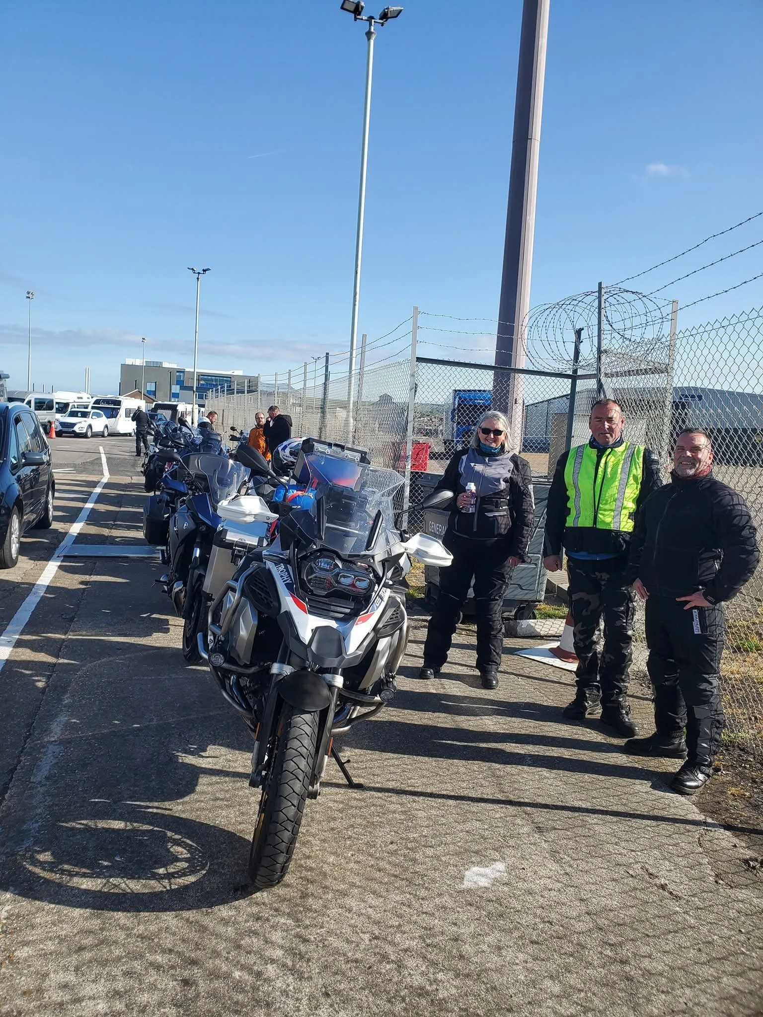 Group of motorcyclists, including three people standing near parked motorcycles and cars, at an outdoor area with a fence, a sky with some clouds, and tall light poles.