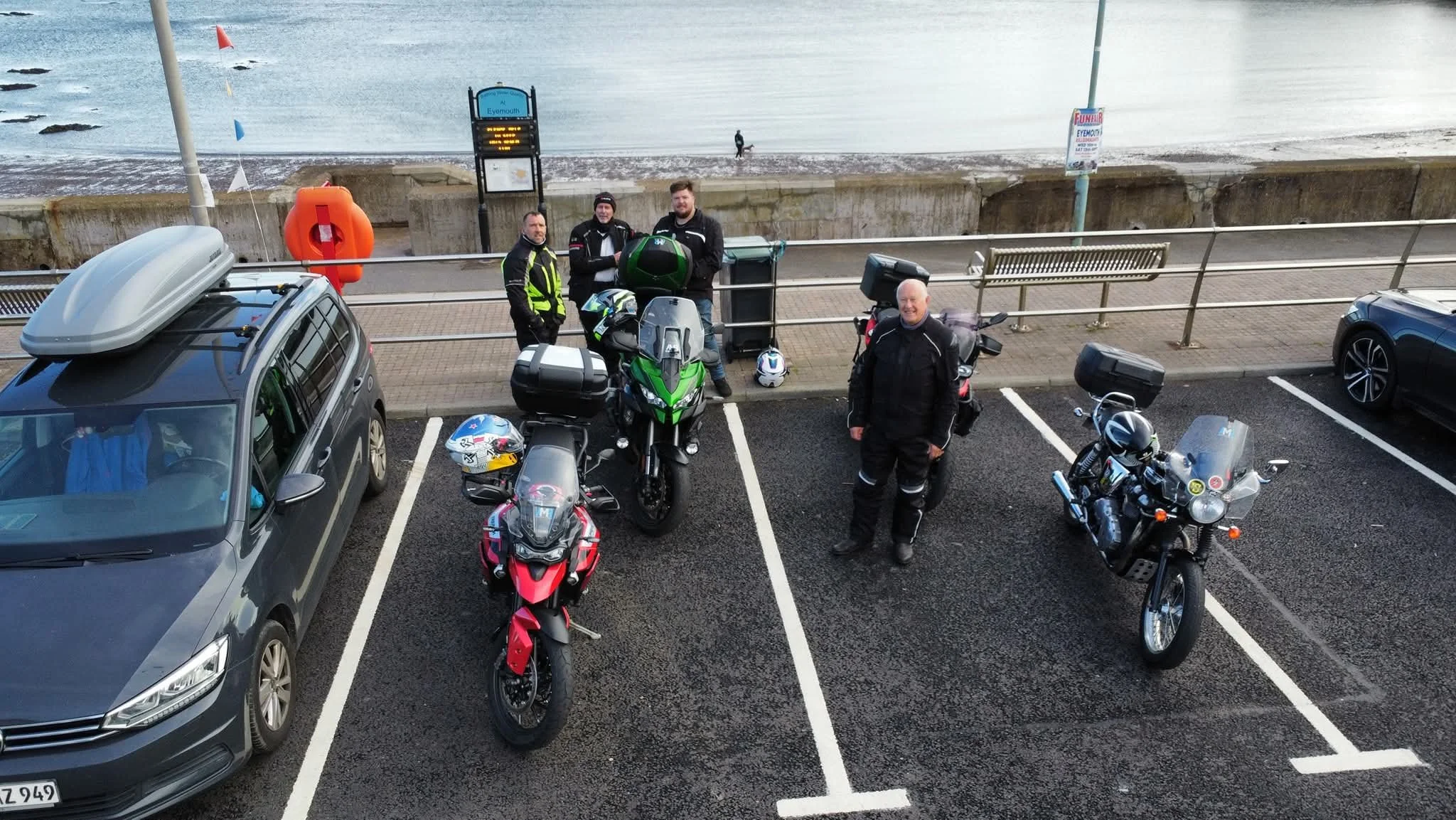 Four motorcyclists standing next to their parked motorcycles and a car on a seaside parking lot, with the ocean in the background.