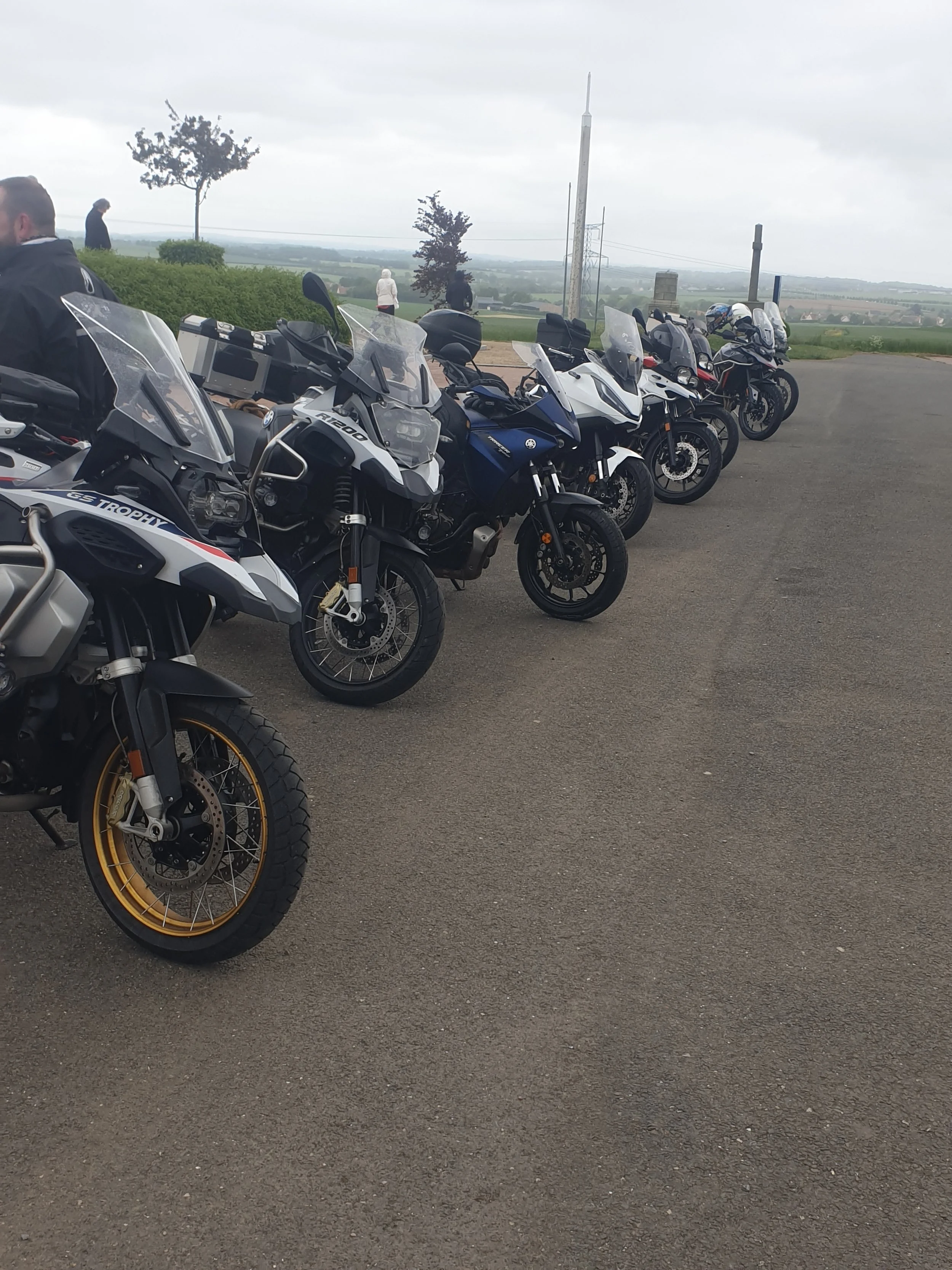 Multiple adventure and touring motorcycles parked on a paved road with a scenic countryside view in the background, under cloudy skies.