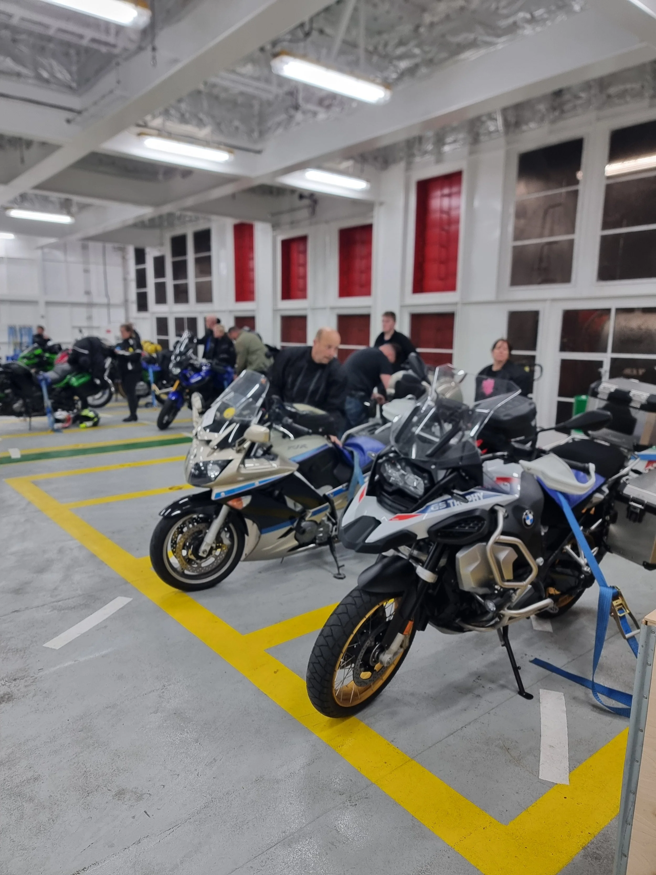 Indoor parking garage with several motorcycles parked, including BMW and Yamaha models, and people standing nearby.