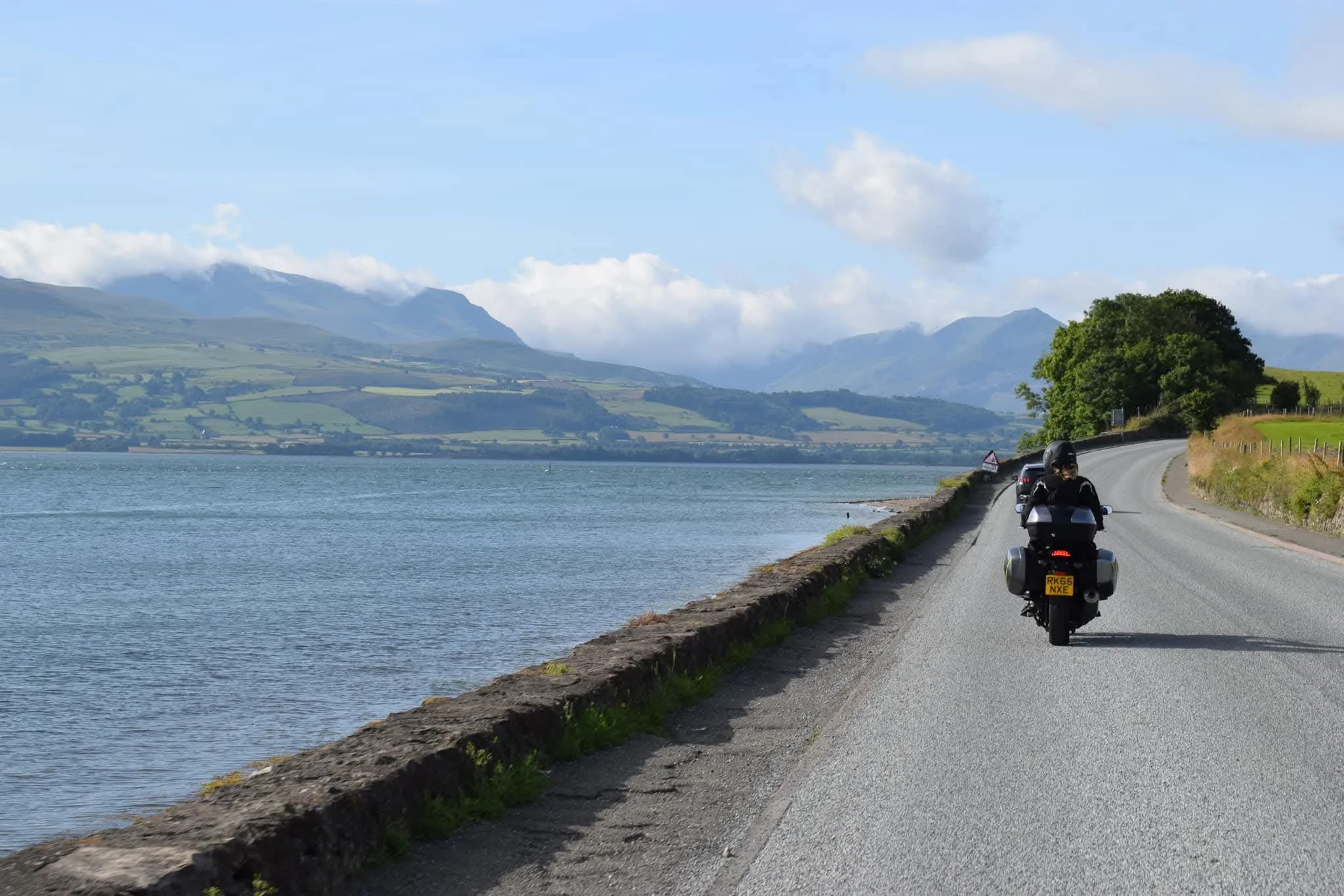 Motorcycle and cars driving along a scenic lakeside road with mountains in the background.
