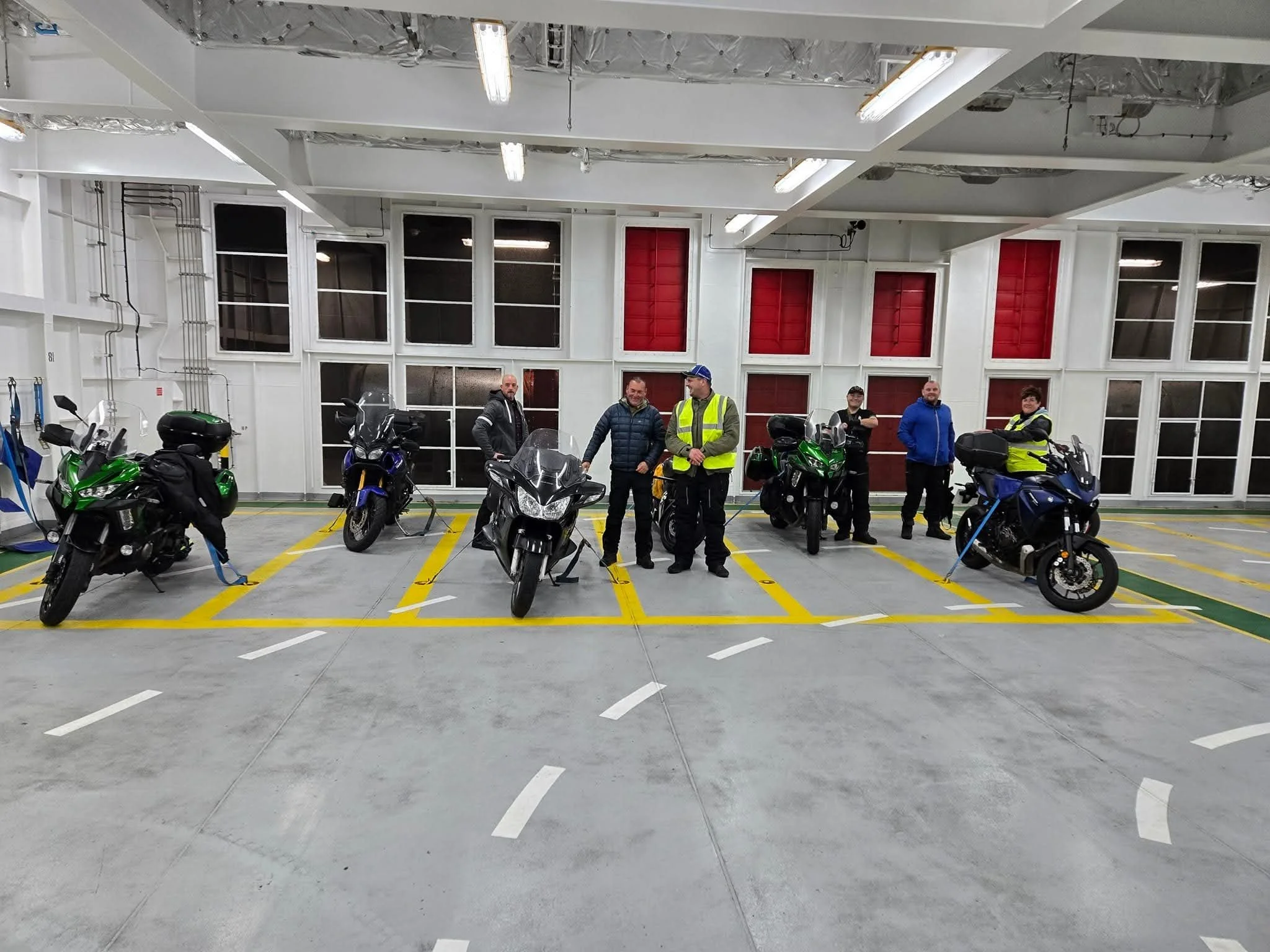 Indoor parking garage with six motorcycles parked in a row and seven men standing behind them, some in jackets and high-visibility vests, against a white wall with large windows and red panels.