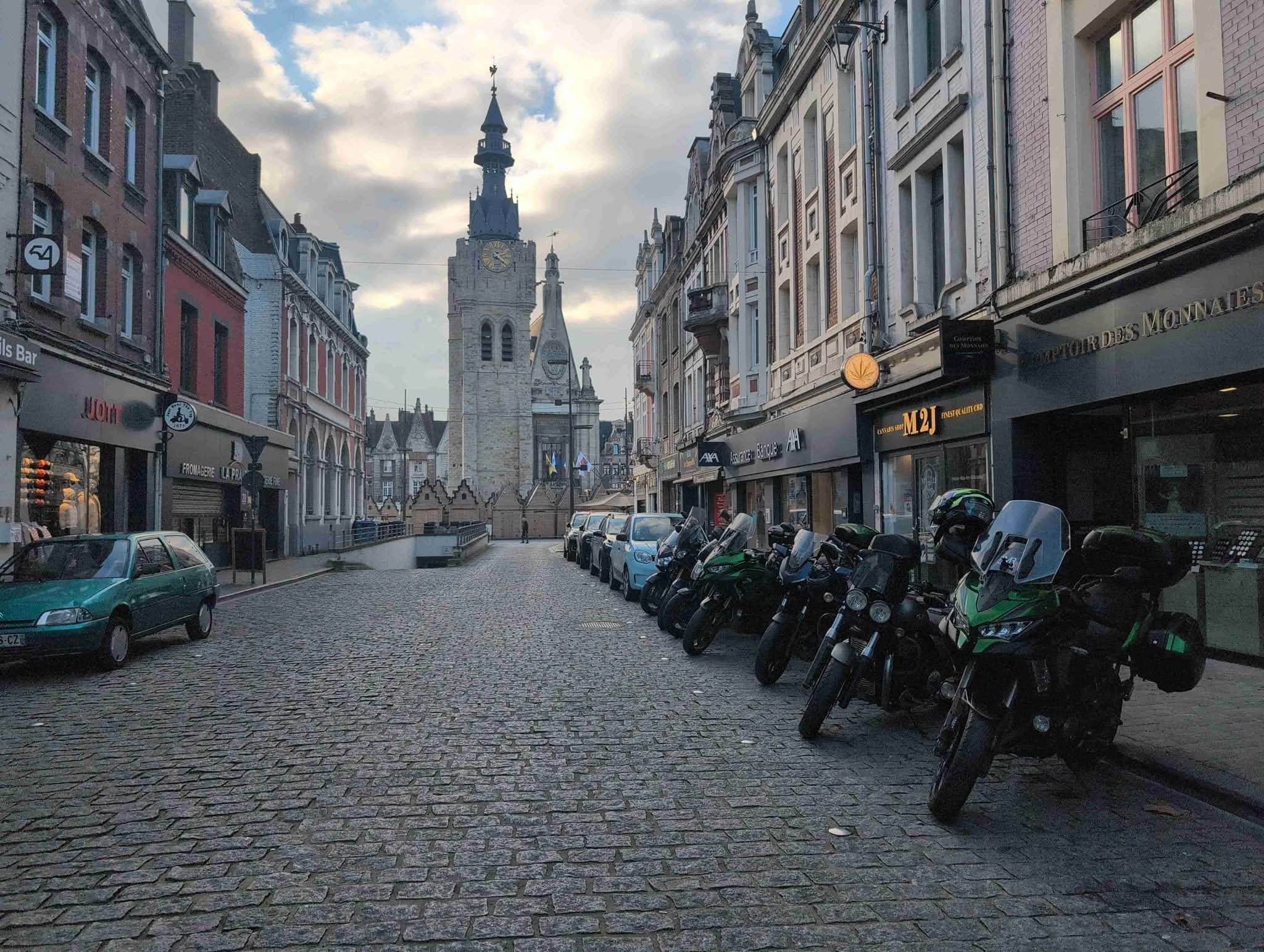 A cobblestone street lined with parked cars and motorcycles, with historic buildings and a tall clock tower in the background under a cloudy sky.