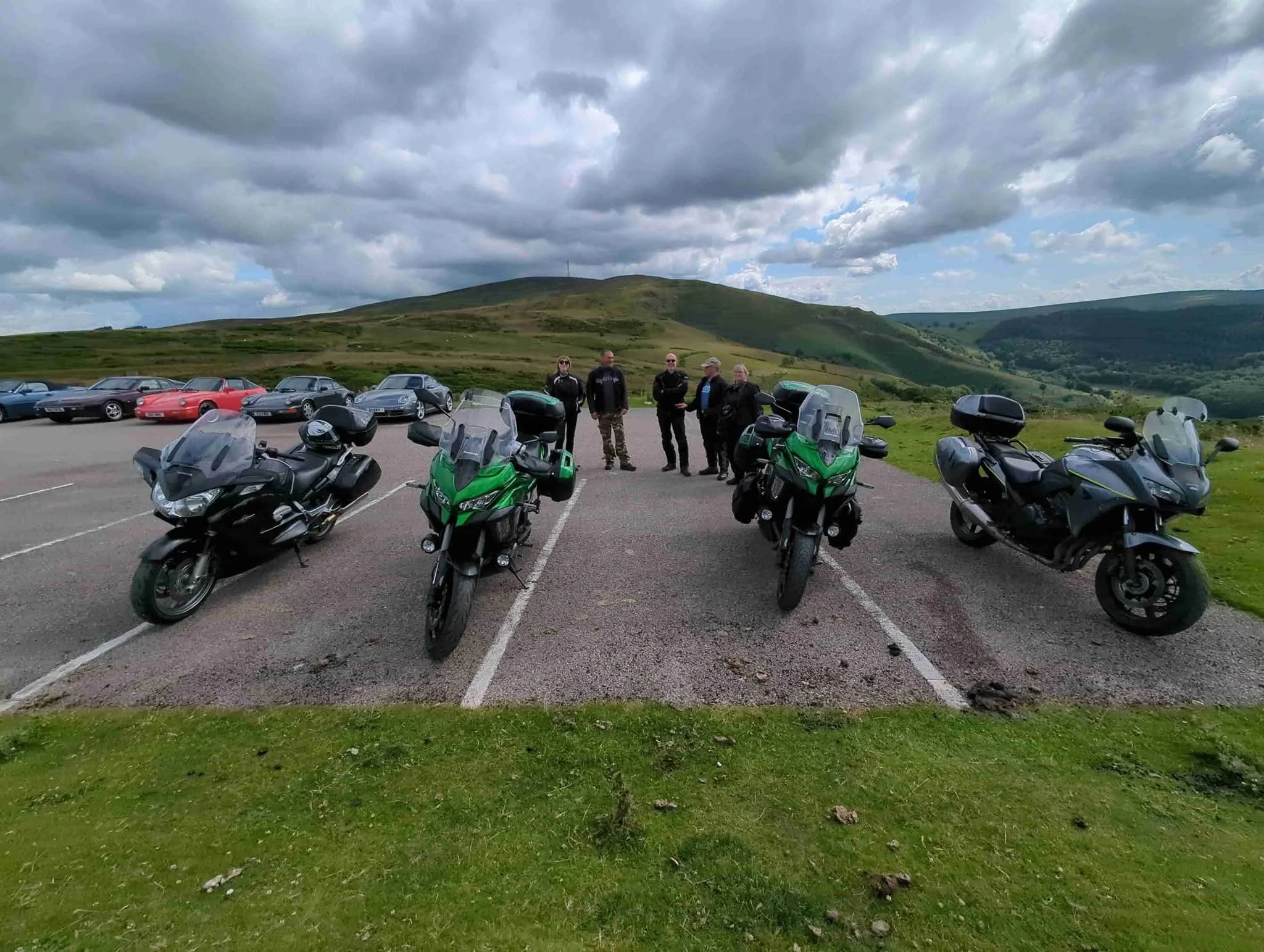 Four motorcycles parked in a lot with three men standing in the background, hills in the distance, and a cloudy sky overhead.