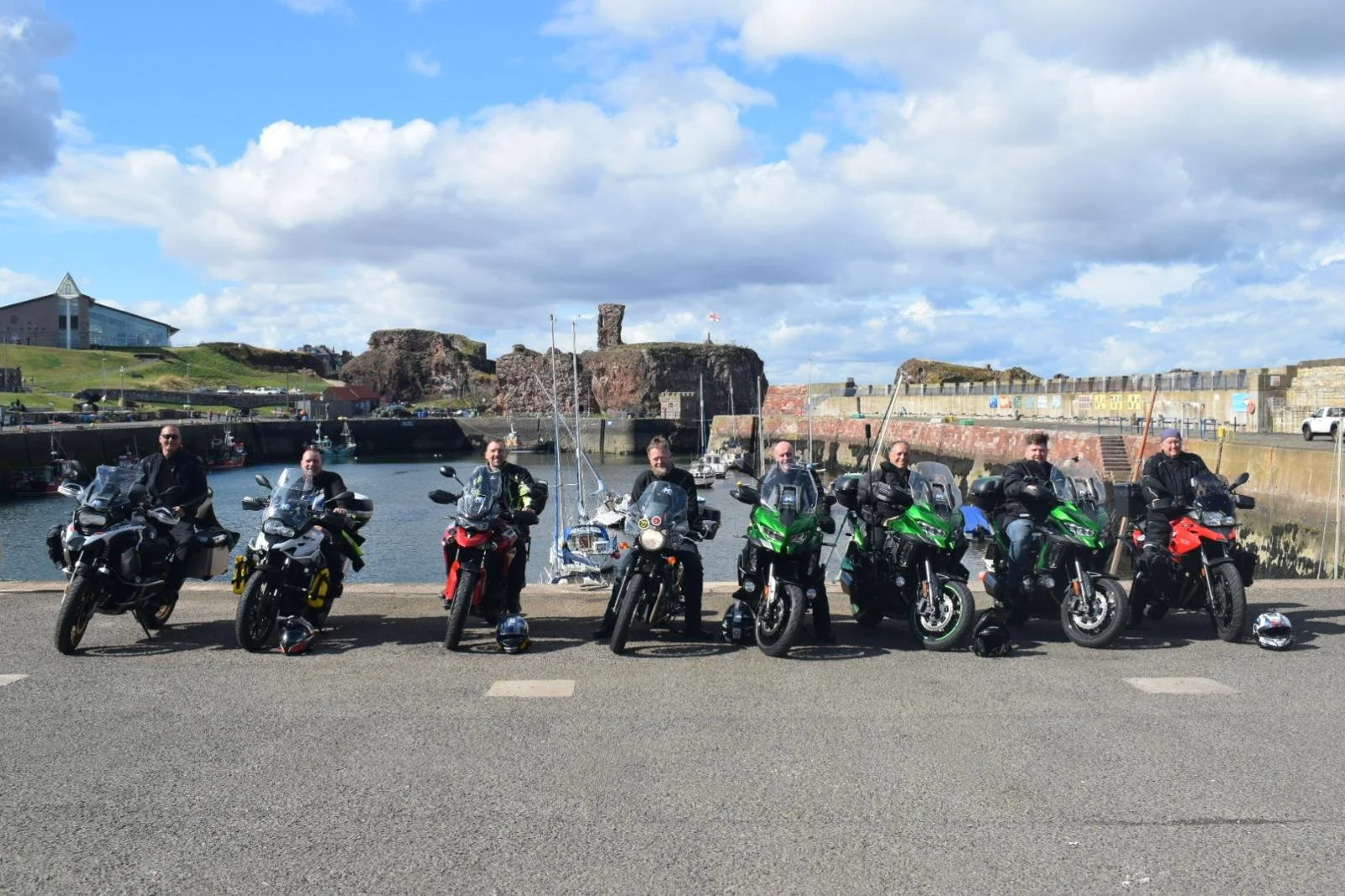 A group of nine motorcyclists on touring bikes parked along a harbor with ships and rocky cliffs in the background under a partly cloudy sky.
