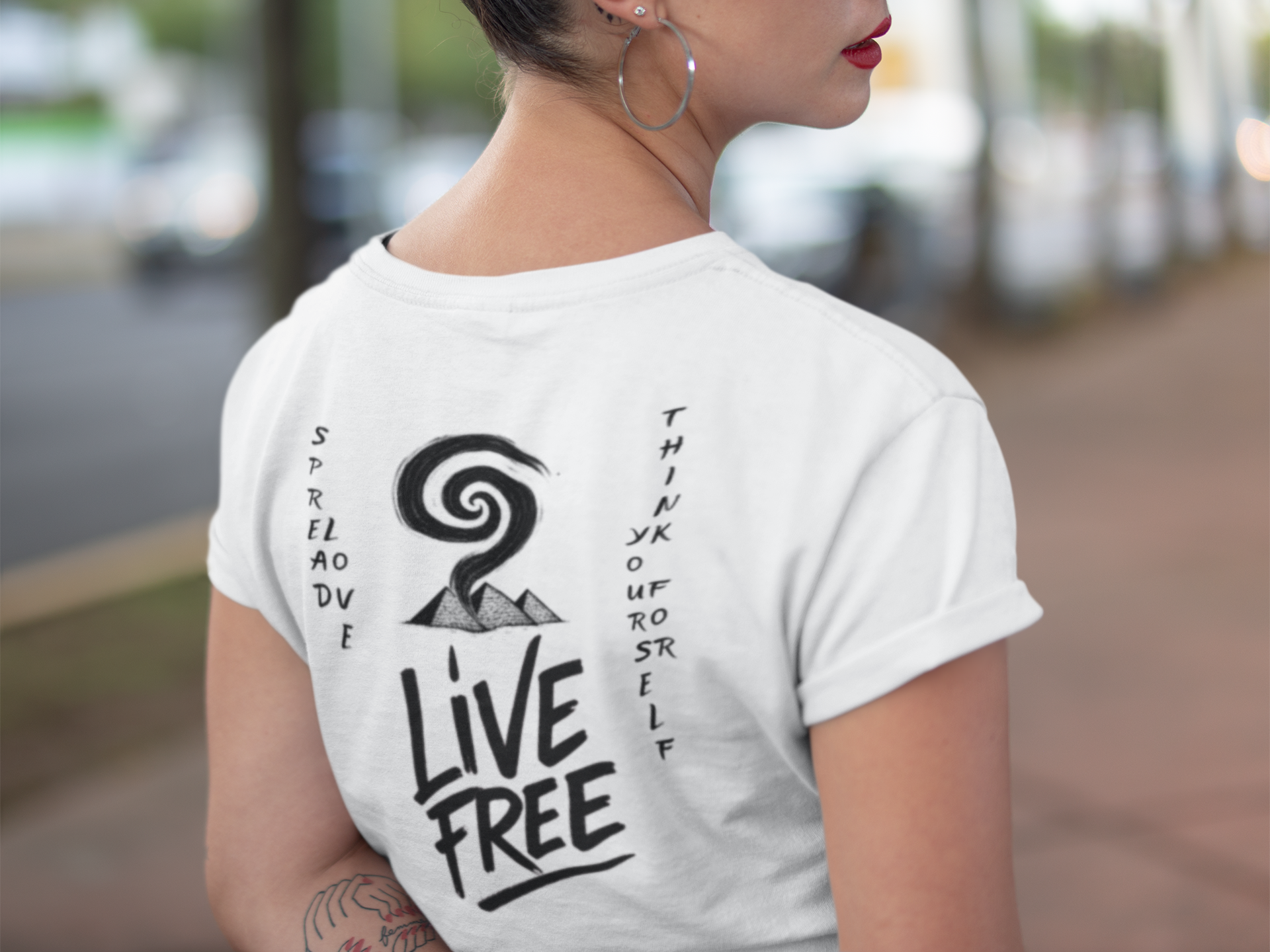 Back view of a woman wearing a white t-shirt with a mountain, spiral smoke, and the words "Live Free" and handwritten phrases about dread and fear. The woman has hoop earrings, red lipstick, and is outside on a sidewalk.