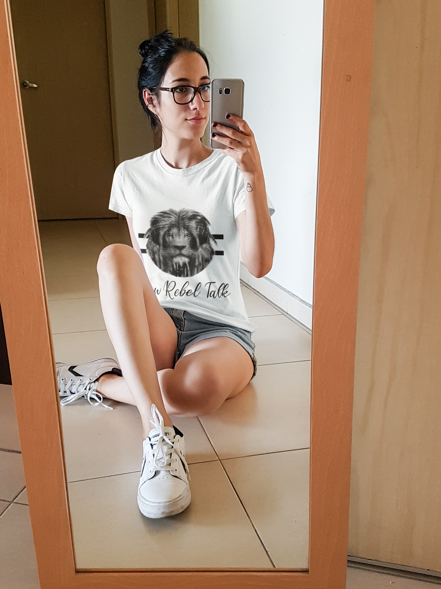 A young woman with dark hair in a bun, wearing glasses, a white t-shirt with a lion graphic and text, gray shorts, and white sneakers, sitting on the floor taking a selfie in a mirror.