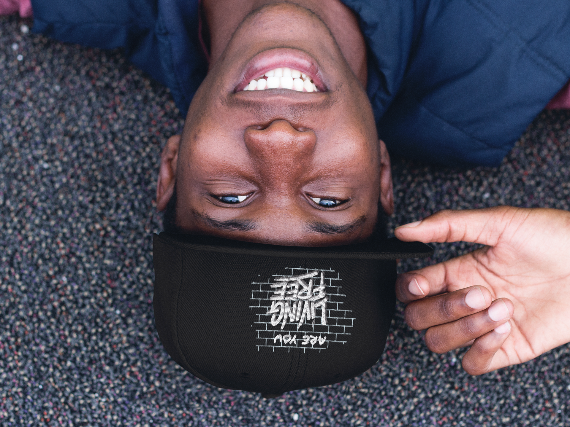 A man lying on multicolored carpet, wearing a black baseball cap with white lettering and a dark jacket, smiling with his mouth open, while someone adjusts his cap.
