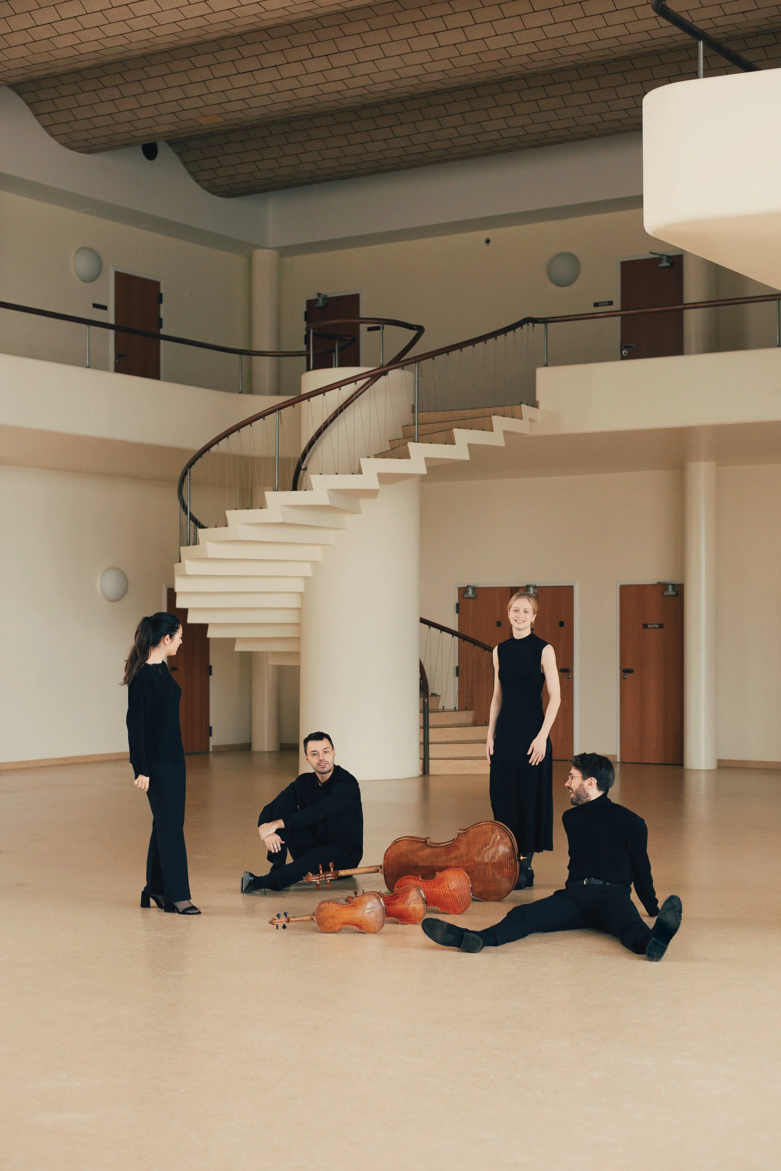 The four members of NOVO Quartet in black clothing in a large, empty room with a curved staircase and wooden doors, two sitting on the floor and two standing, with cello instruments on the floor.