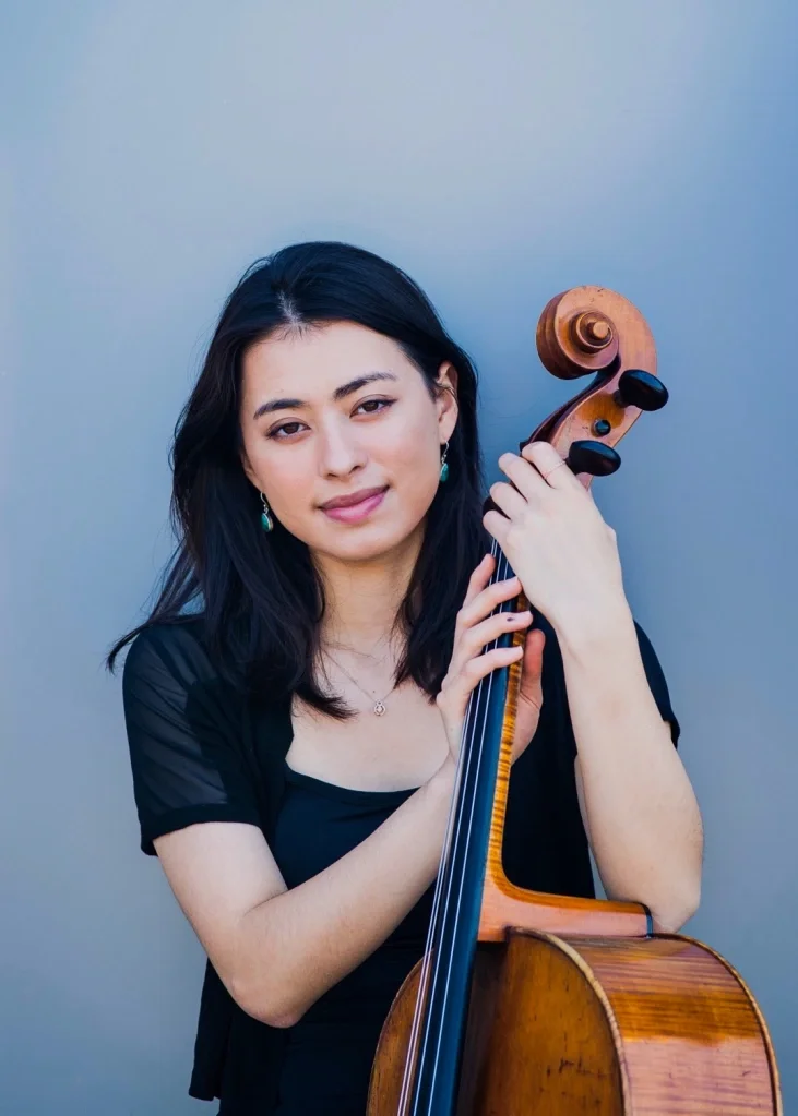 Riya Hamie sits in a black dress, holding a cello against a gray background.