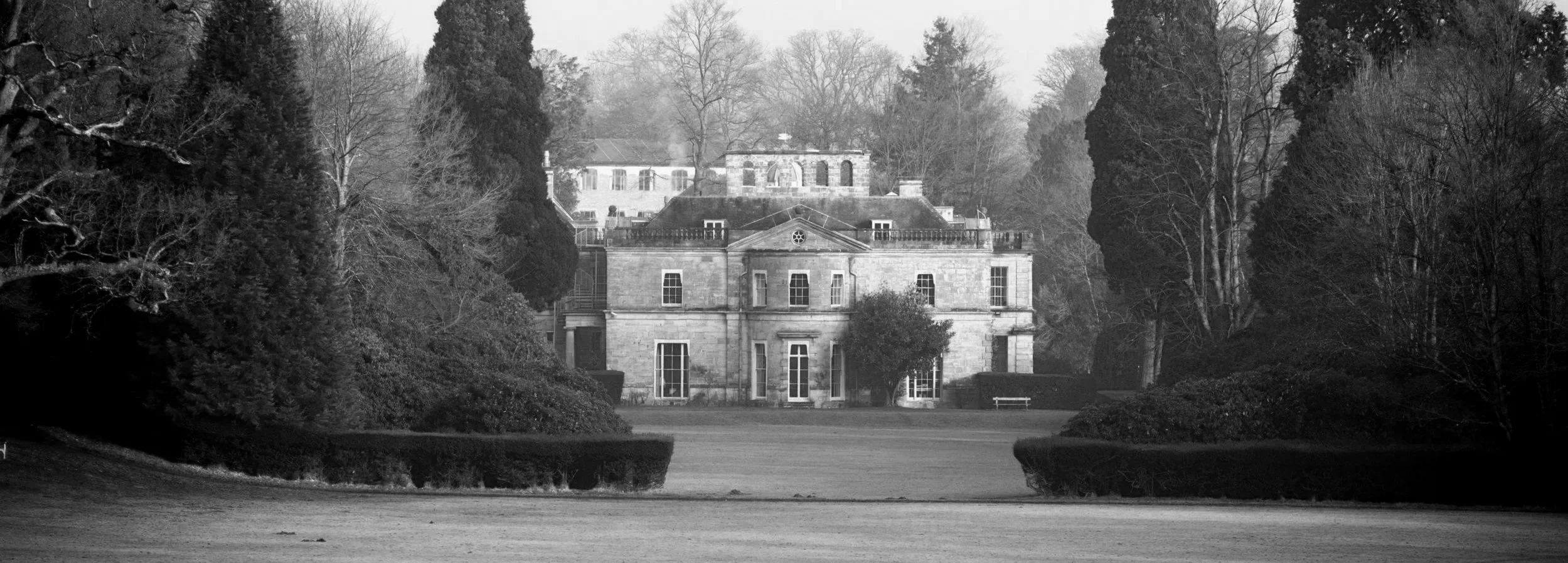 Kidbrooke Mansion: A large historic mansion with multiple stories, arched windows, and a central entrance, surrounded by tall trees and well-maintained gardens, seen in black and white.