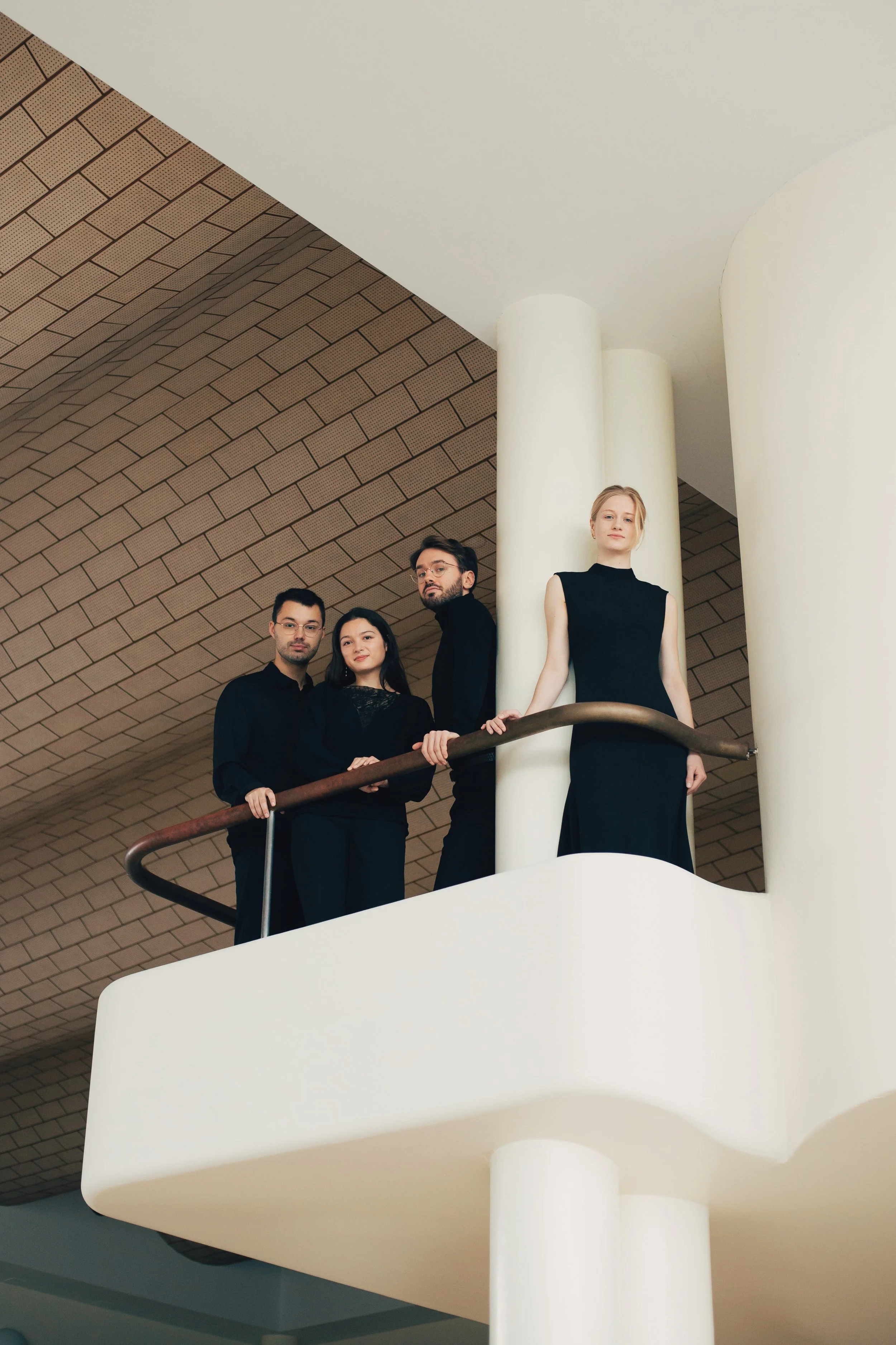 The four members of NOVO Quartet in black standing on a white balcony with brick walls and large cylindrical columns.