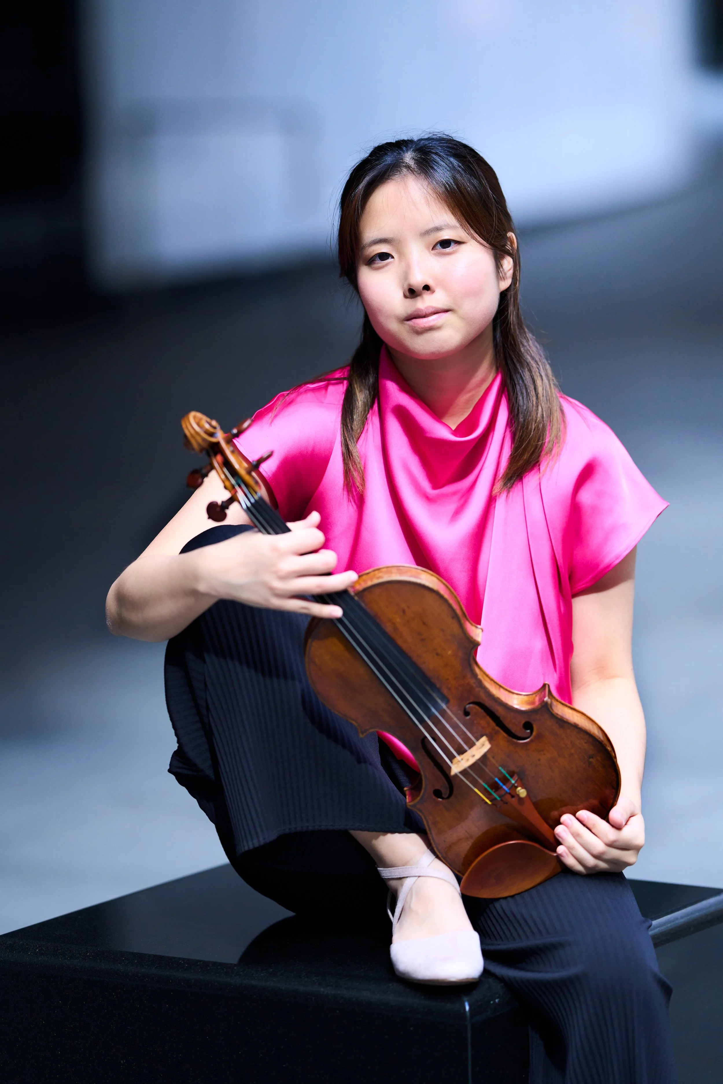Hana Chang sitting on a black platform holding a violin, wearing a vibrant pink satin top.