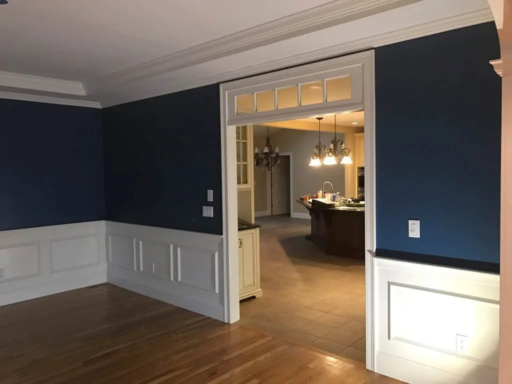 View of a dining room with hardwood floors, white wainscoting walls, and a doorway leading to a kitchen area with hanging light fixtures, a kitchen island, and beige tiled flooring.