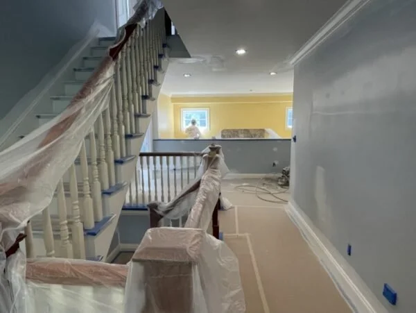 Interior of a house under renovation with protective coverings on stairs and furniture. The room is painted yellow with large windows allowing natural light.