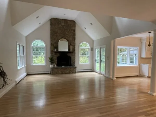 Empty living room with hardwood floors, a stone fireplace with a mirror, and large windows with greenery outside.