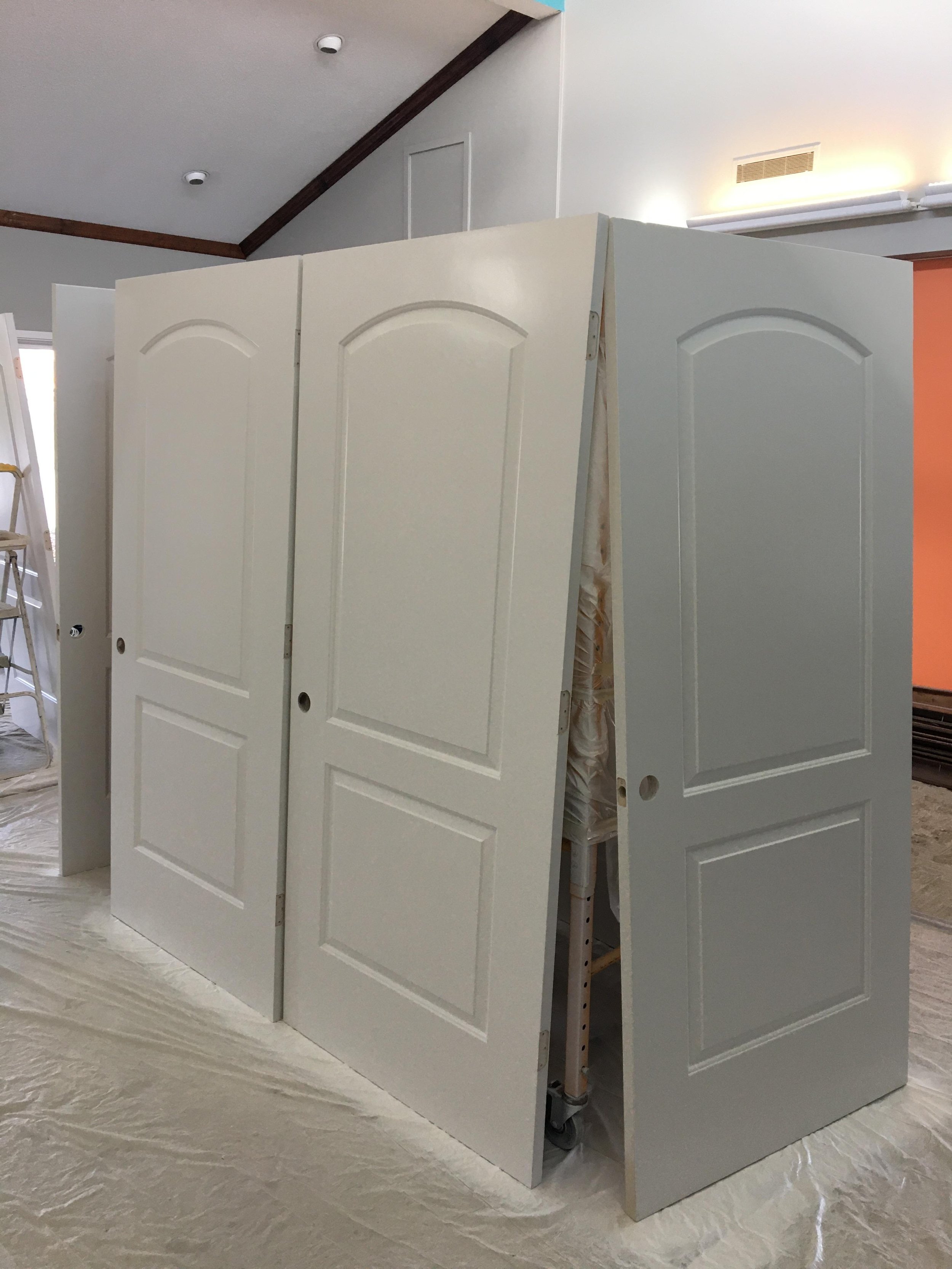 A set of four white interior closet doors leaning against each other in a room, with a ladder and a ladder to the left and some construction materials and a vent on the wall in the background.