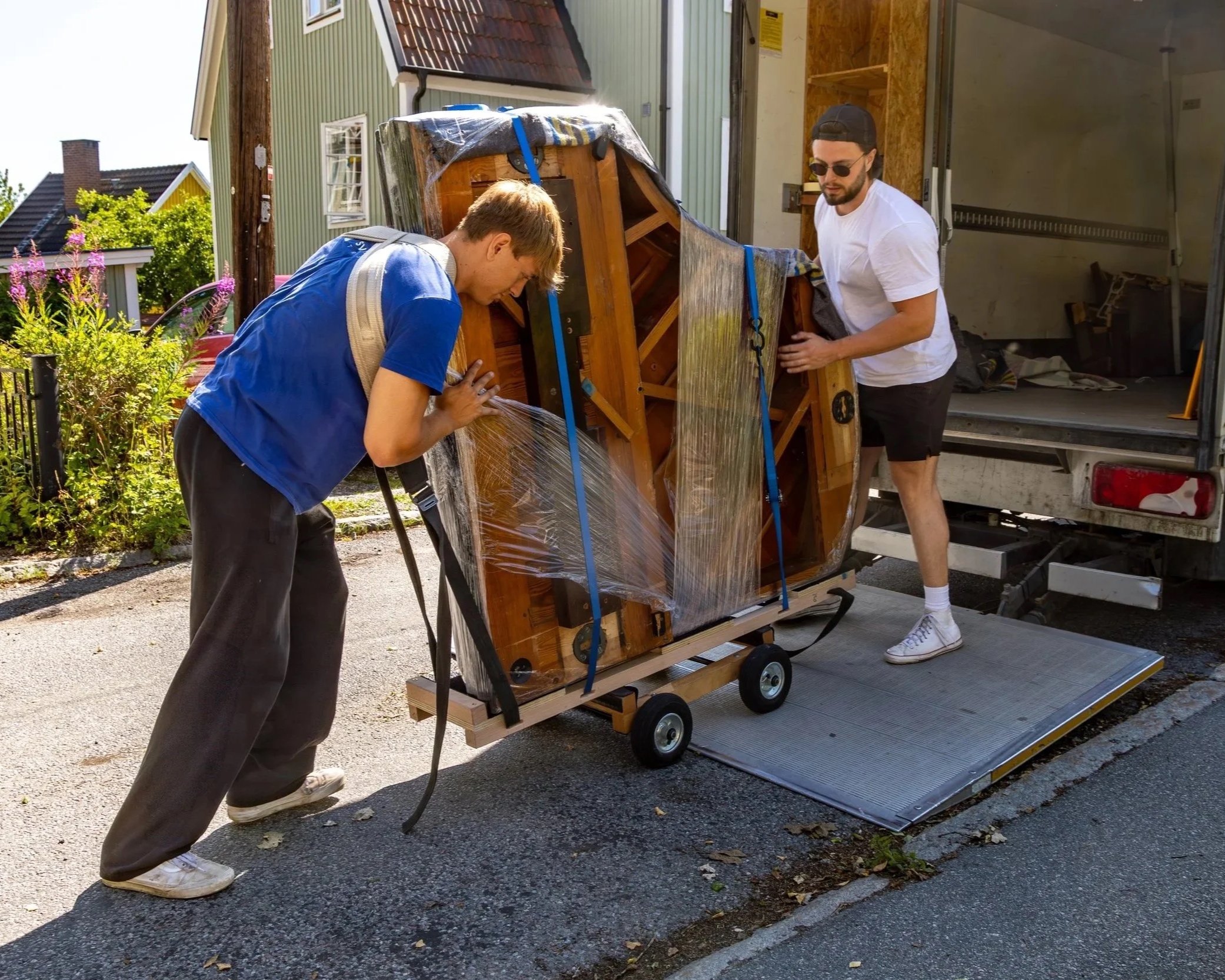 Two men are loading a wooden furniture piece wrapped in plastic onto a moving truck outside a house. One man wears a blue shirt and the other a white shirt and black shorts, both are guiding the furniture onto a ramp.
