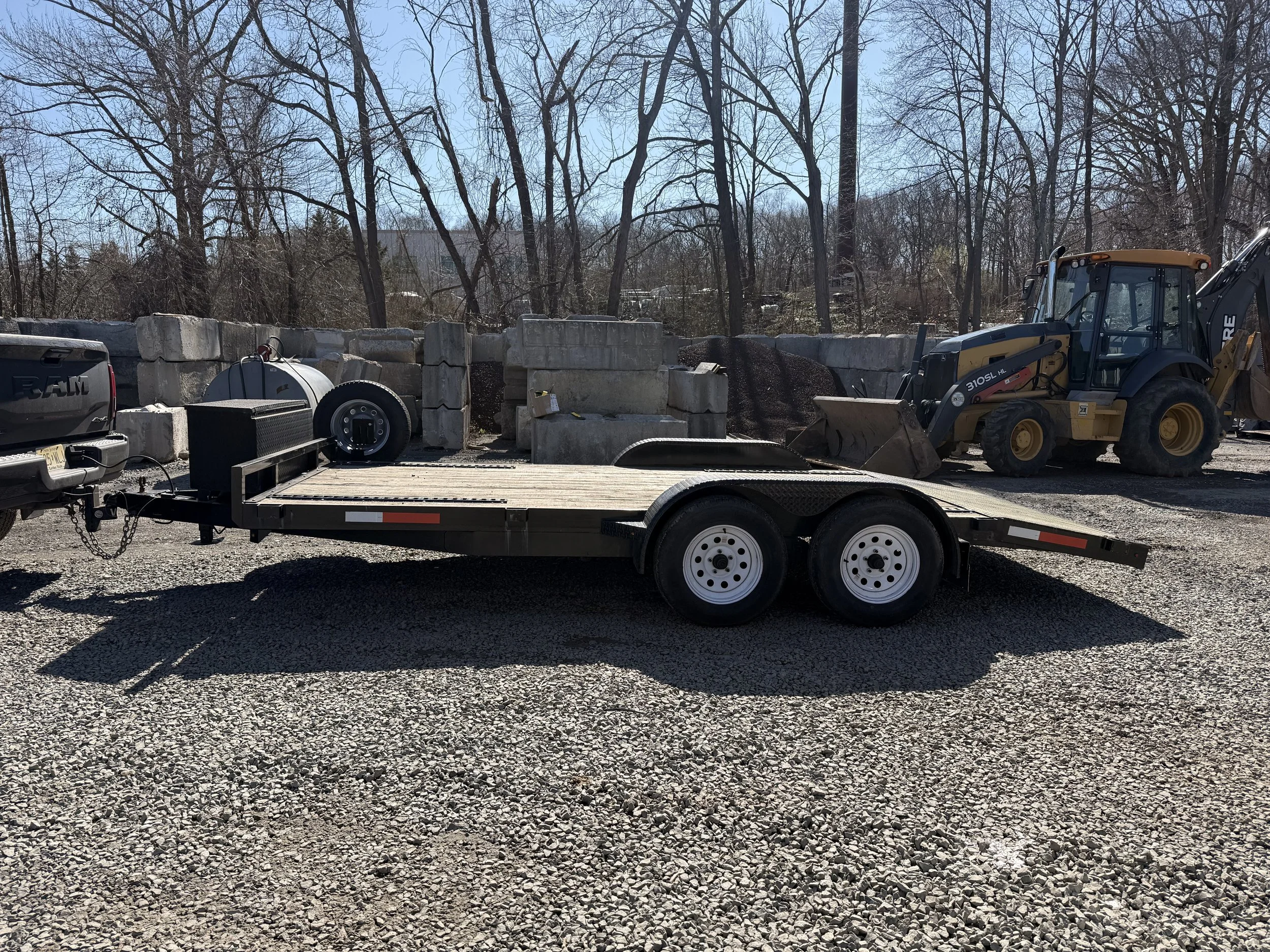 Flatbed trailer parked on gravel lot with construction equipment and concrete blocks in background.