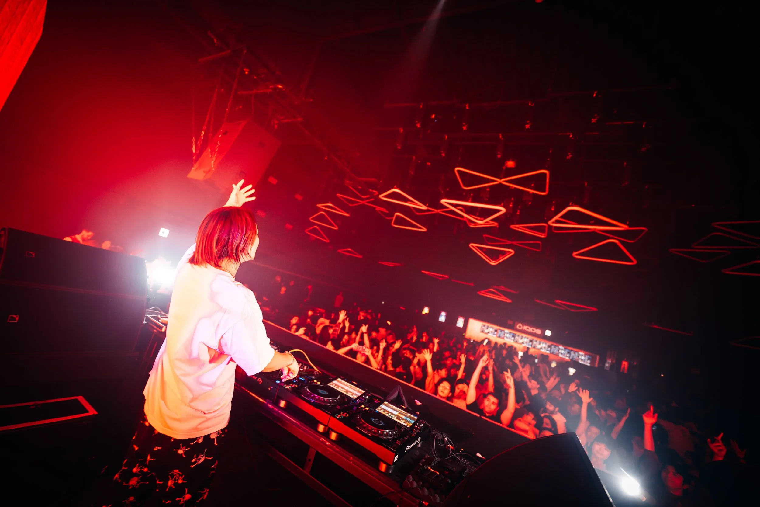 A female DJ with red hair performing at a nightclub, facing a crowd with arms raised, illuminated by red lighting and geometric light fixtures hanging from the ceiling.